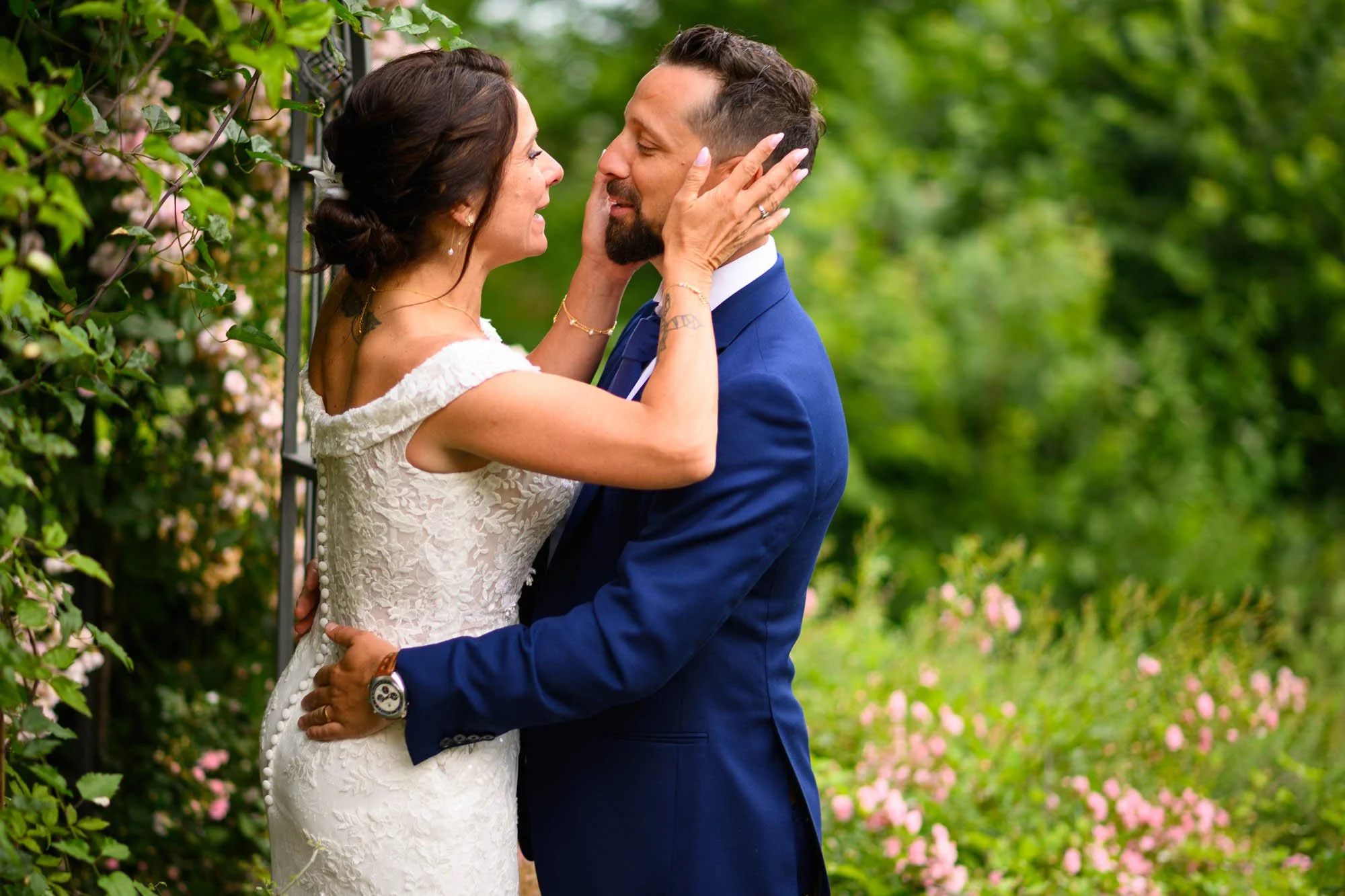 Un couple de mariage se regarde dans un jardin, lui en costume bleu et elle en robe blanche, elle le tient de face et lui caresse le visage.