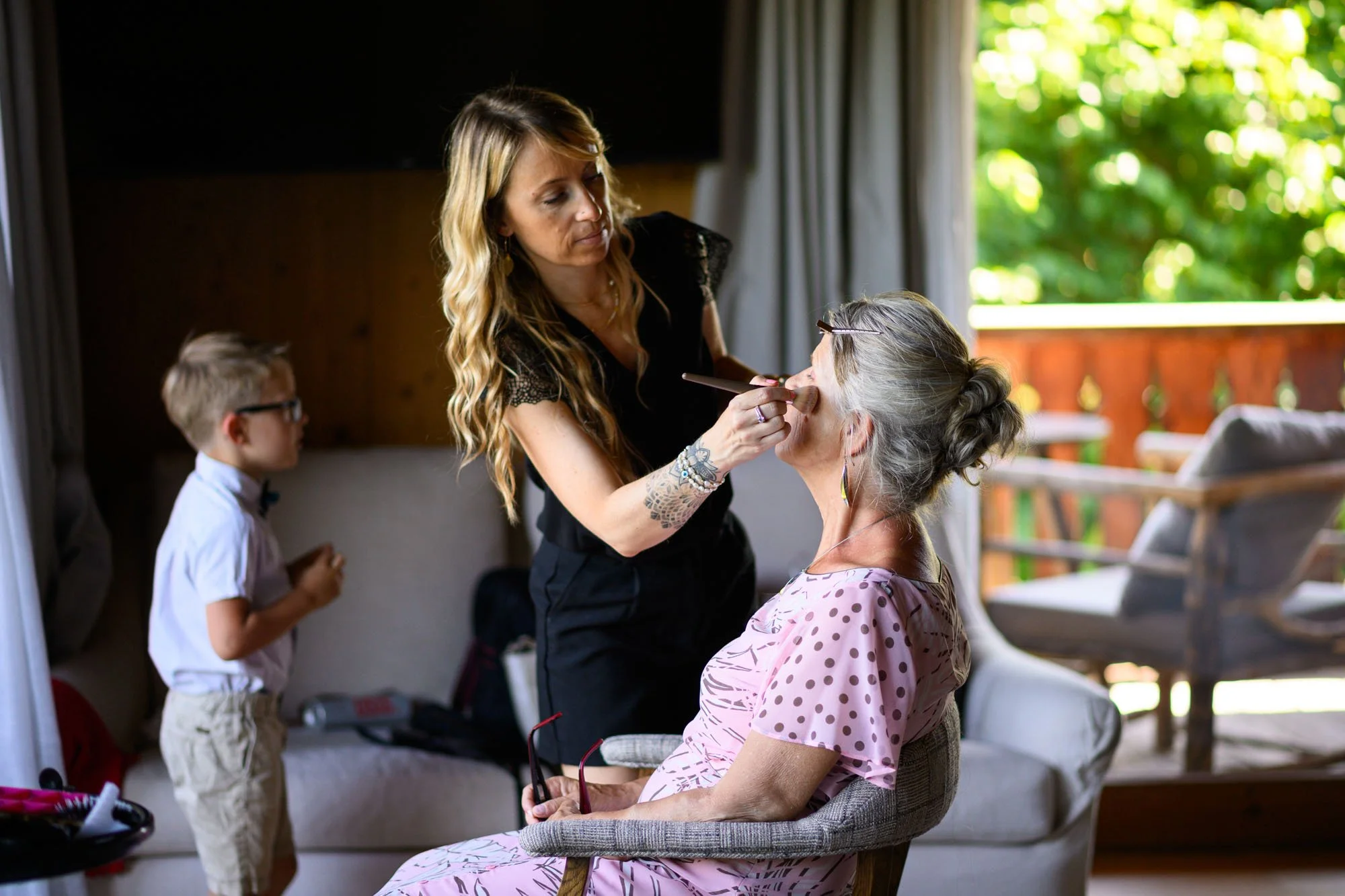 Une femme fait du maquillage à une femme âgée assise, dans un environnement intérieur avec balcon et vue sur la nature.