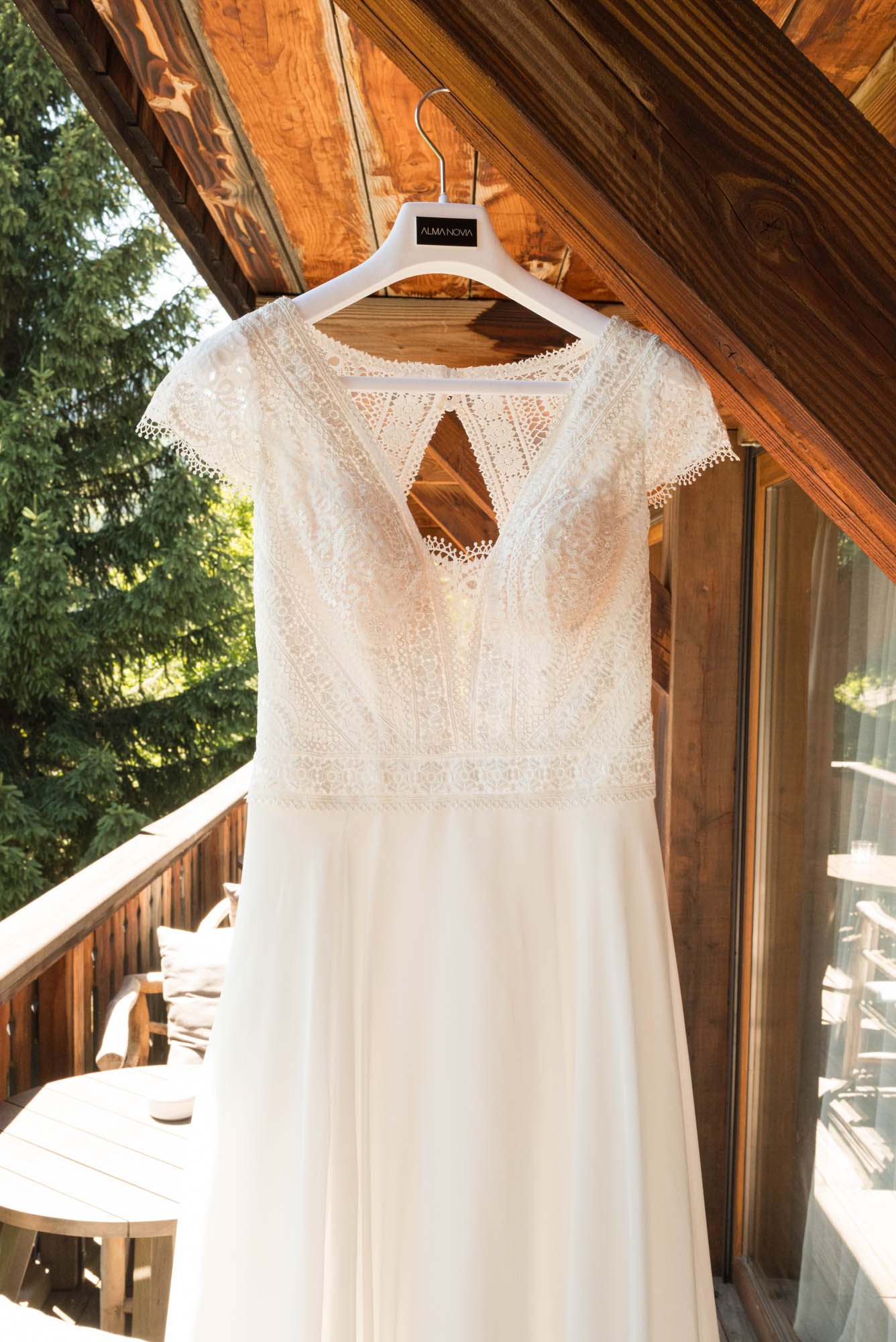 Robe de mariage blanche avec dentelle, suspendue à une porte de balcon en bois, avec vue sur des arbres verts.