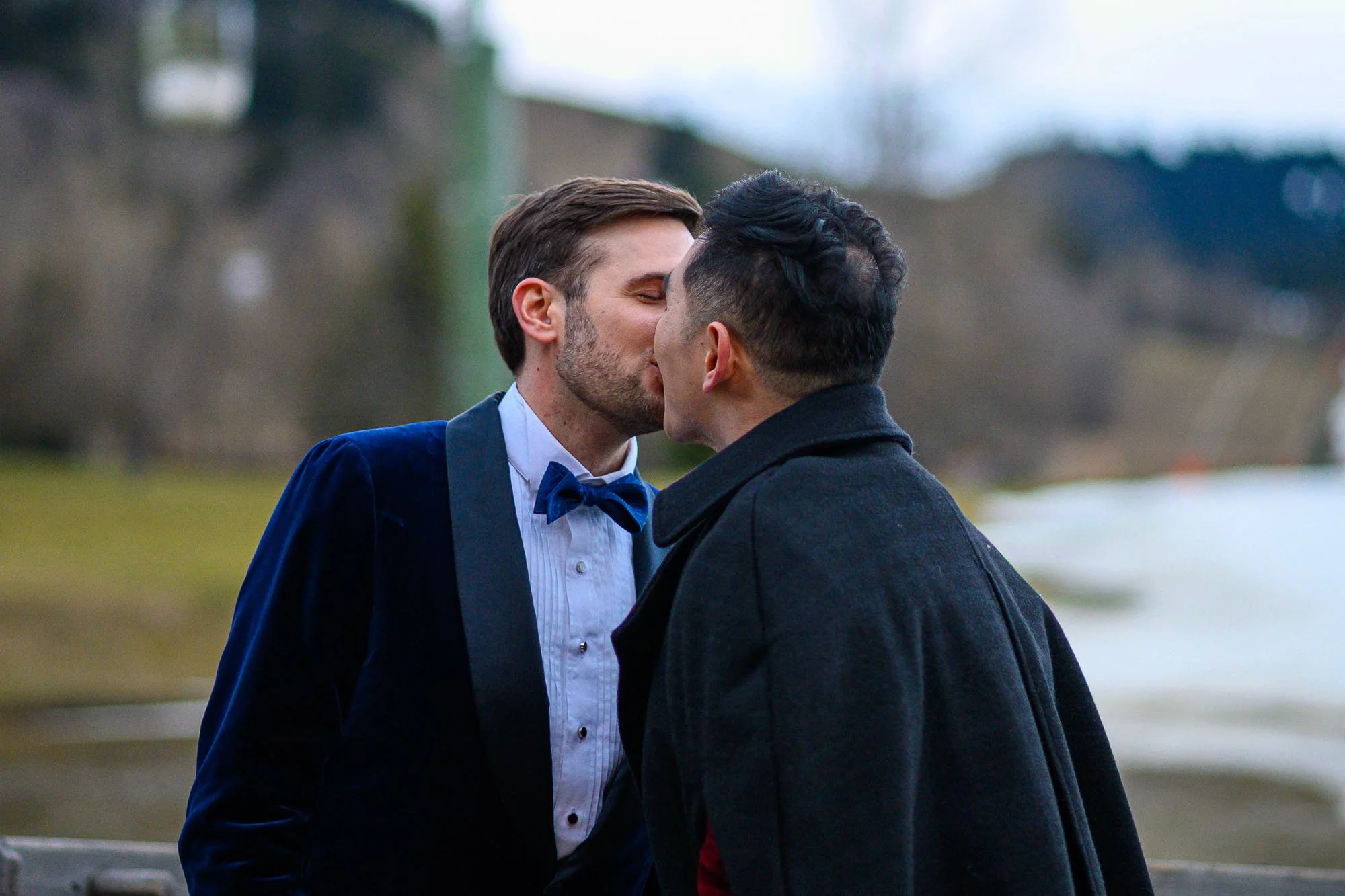 Deux hommes s'embrassent et échangent un baiser dans une ambiance romantique en plein air, avec un arrière-plan flou de paysage.
