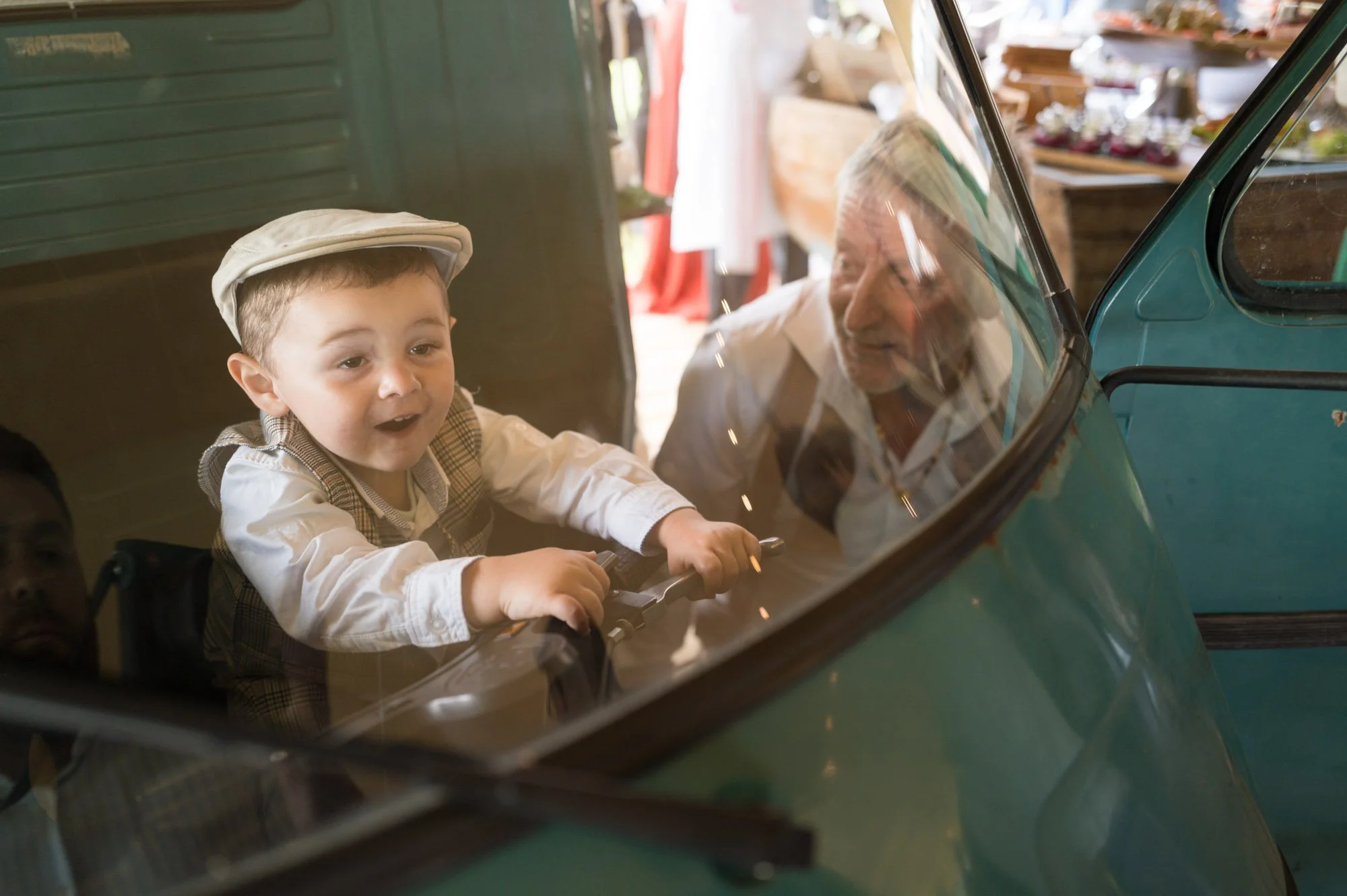 Un jeune garçon avec une casquette et des vêtements d'époque à l'intérieur d'un véhicule classique vert, avec un homme plus âgé regardant à travers la fenêtre.