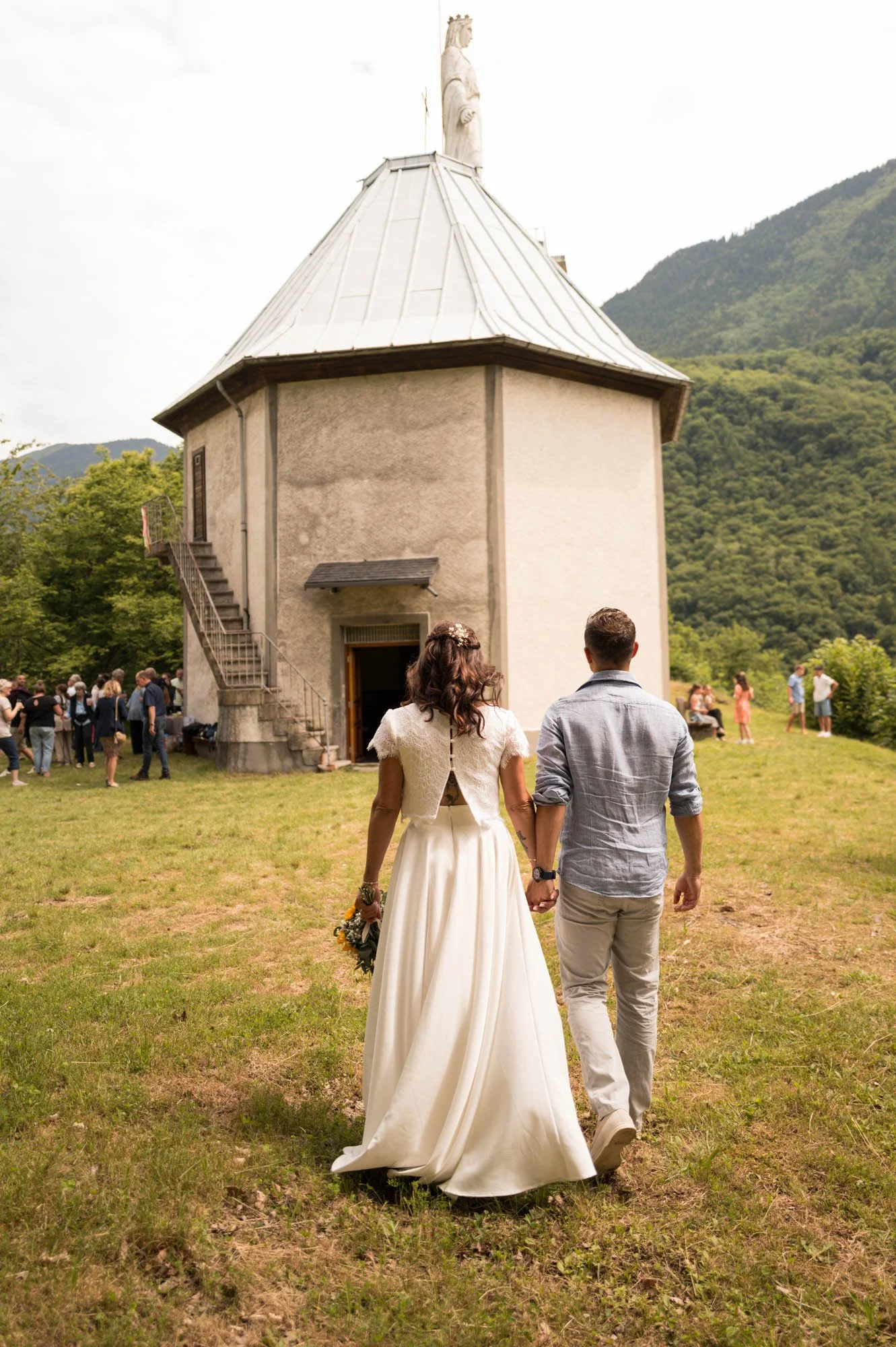 Un couple marié marche vers une chapelle au fond d'une pelouse verte lors d'une cérémonie de mariage avec plusieurs invités.