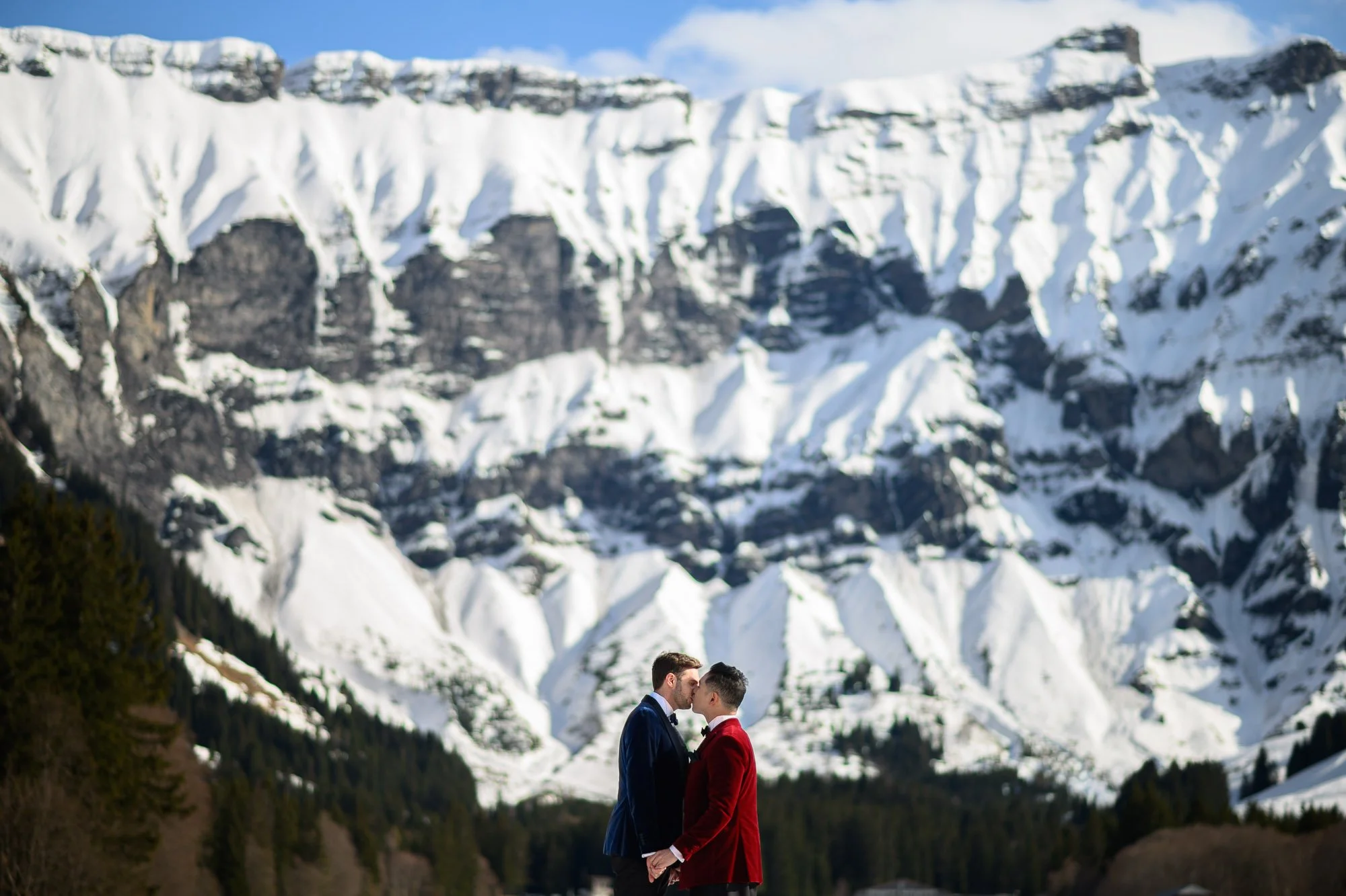 Deux hommes habillés en costumes formels, un en bleu et l'autre en rouge, se tenant face à face, les yeux fermés, devant un paysage de montagnes enneigées.