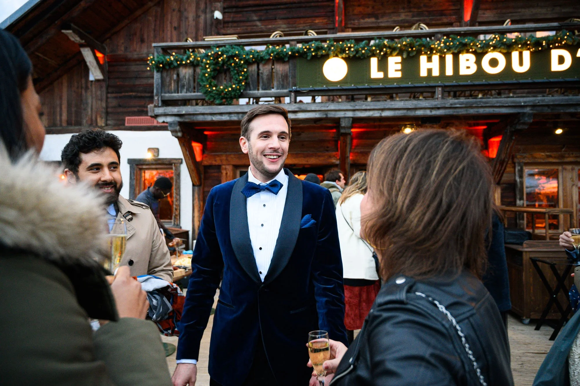 Groupe de personnes lors d'une fête ou d'un événement à l'intérieur d'une cabane en bois décorée avec des guirlandes lumineuses, en train de parler et de boire du champagne.
