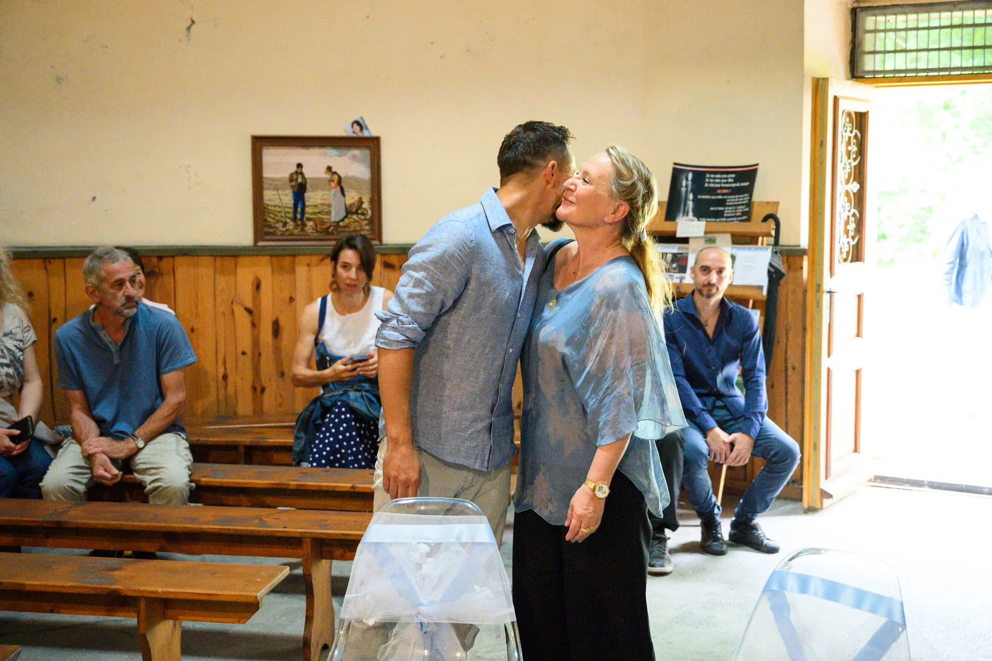 Un homme et une femme s'embrassent dans une pièce, entourés de plusieurs personnes assises sur des bancs en bois, dans un cadre intérieur chaleureux.