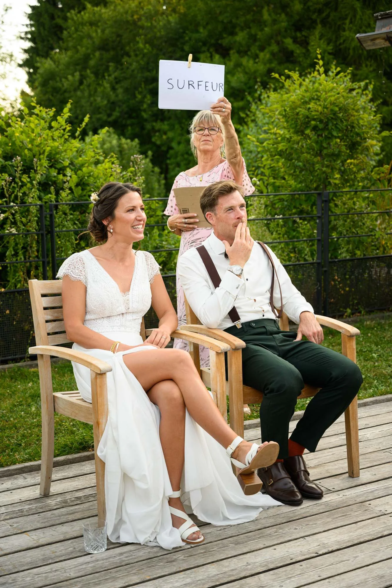 Une femme âgée tient une pancarte avec le mot 'SURFEUR' lors d'un mariage, avec un couple assis en avant d'elle, la femme en robe blanche et l'homme en chemise blanche et pantalon noir.
