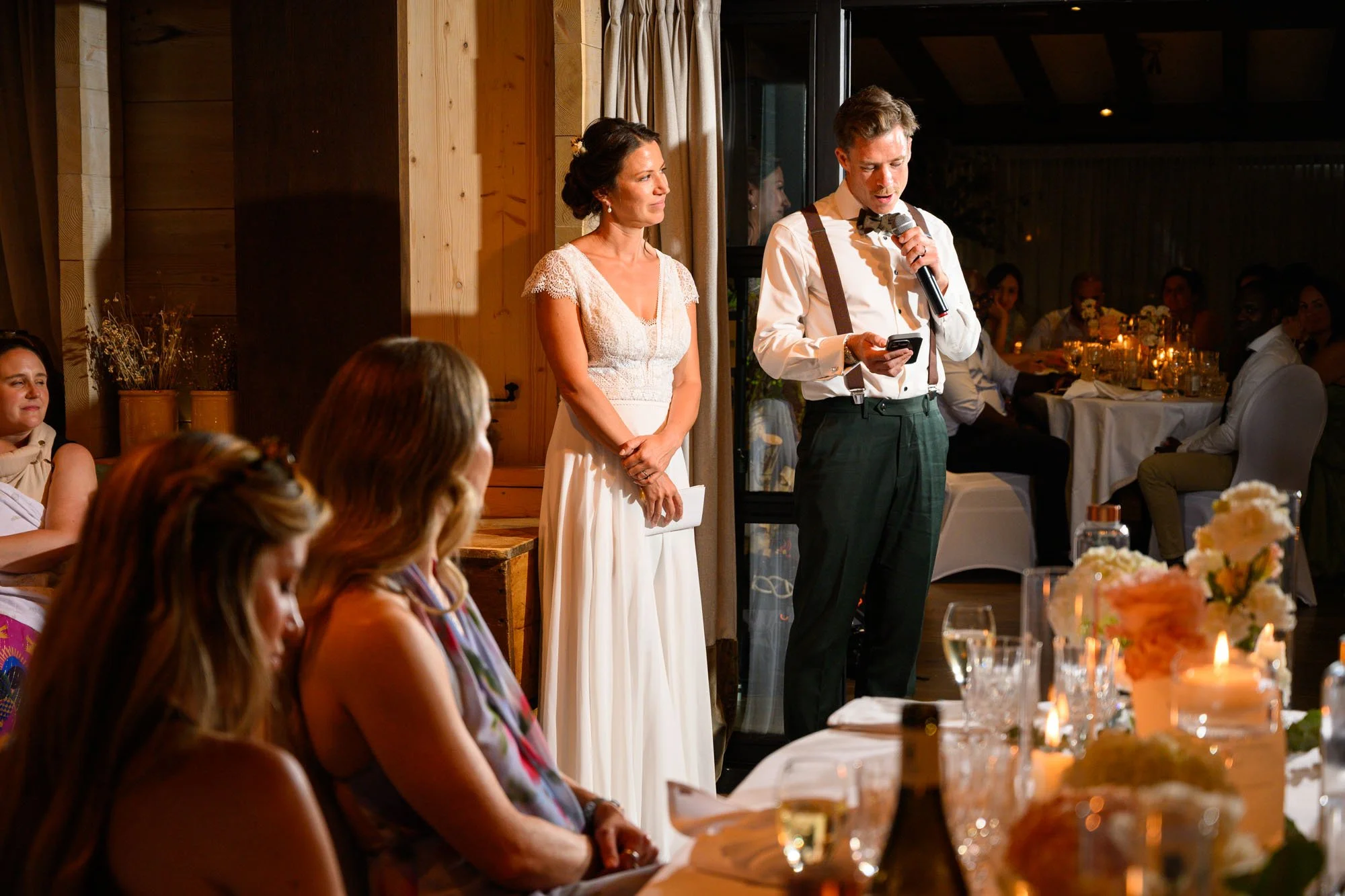 Un discours lors d'un mariage, avec une femme en robe blanche et un homme en chemise blanche et pantalon vert foncé, tenant un micro