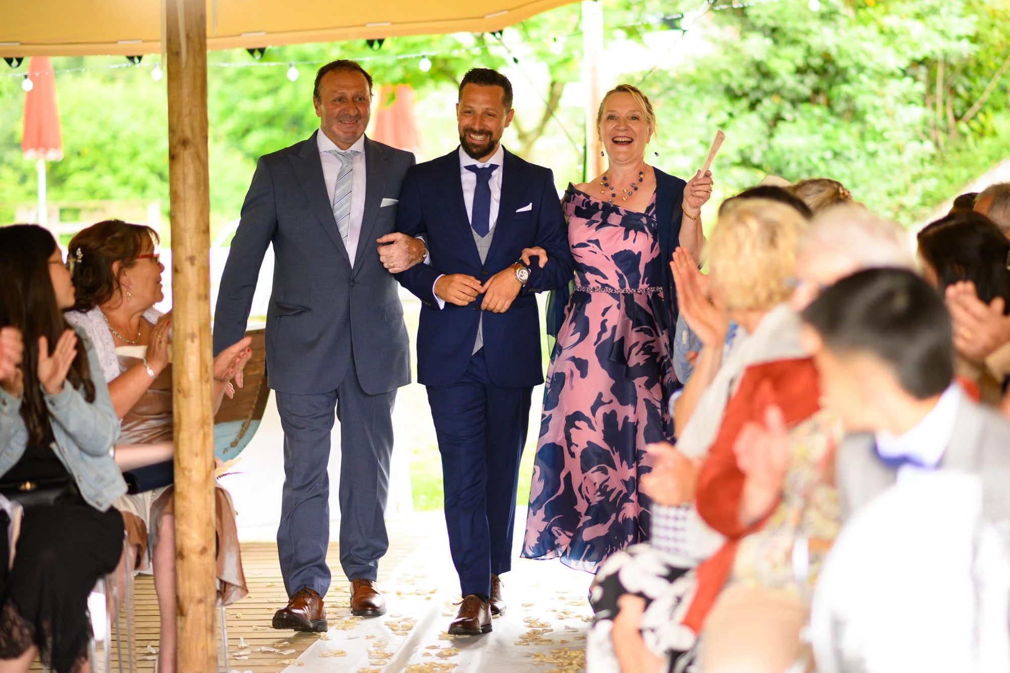 Trois personnes marchent dans une cérémonie de mariage en plein air, entourées d'invités assis et applaudissant. La scène est sous une tente avec un décor naturel verdoyant en arrière-plan.