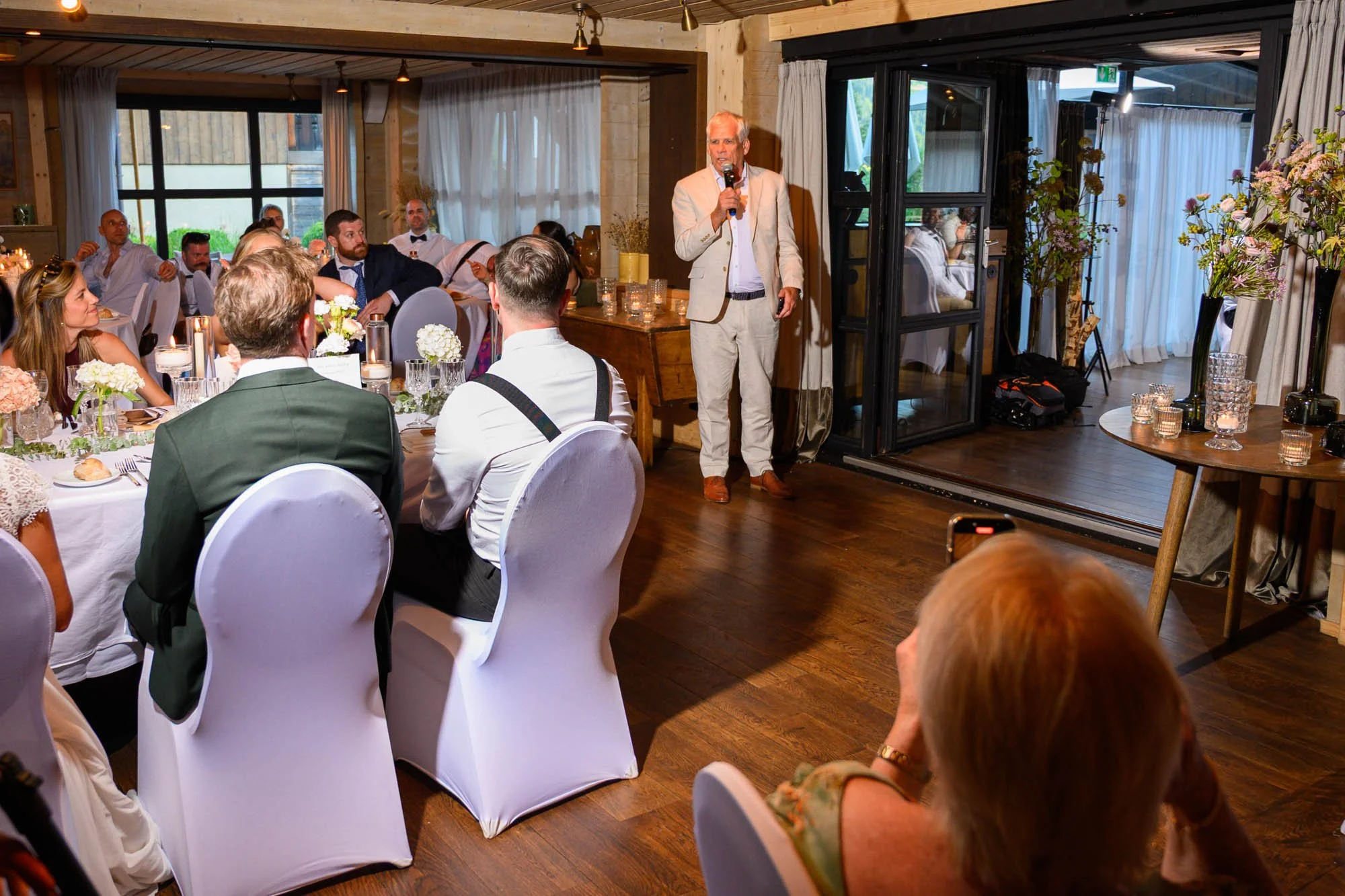 Un homme en costume clair parle avec un micro lors d'un événement organisé dans une salle décorée avec des fleurs et des bougies, face à un groupe de personnes assises à une table.