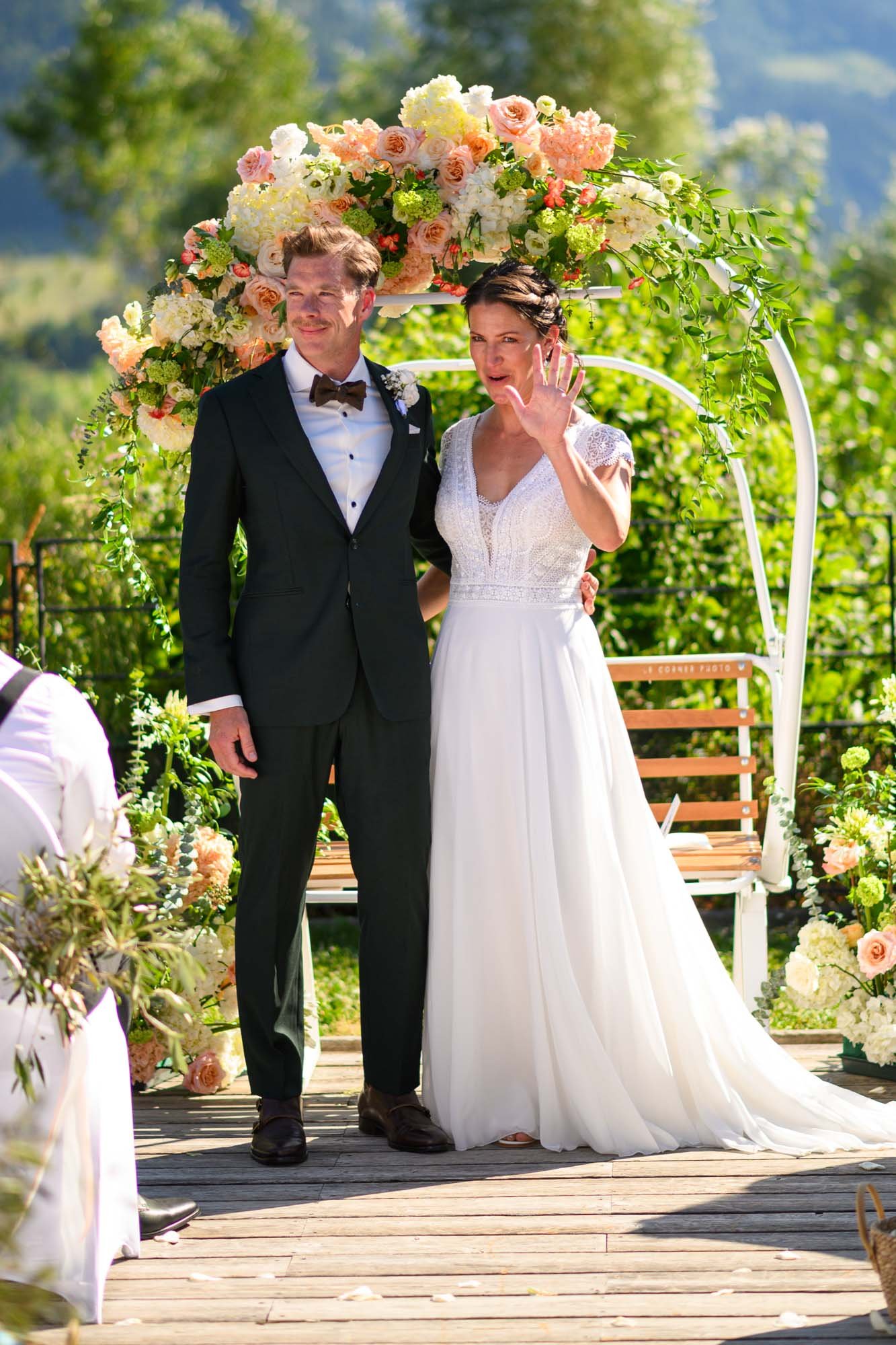 Un couple en vêtements de mariage se tient sous une arche ornée de fleurs, lors d'un mariage en plein air. La femme fait un geste avec sa main, tandis que l'homme la regarde avec un sourire.