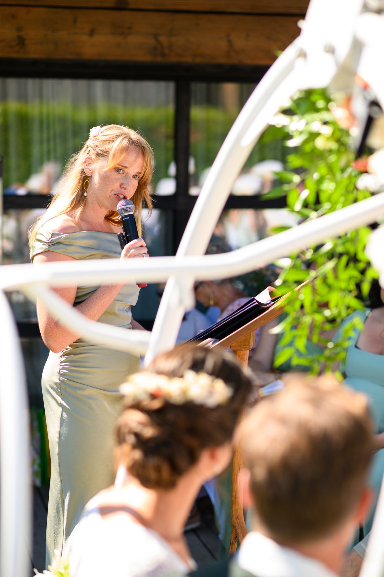 Une femme en robe dorée jaune chante ou parle dans un microphone lors d'une cérémonie en plein air. Il y a des personnes assises en arrière-plan et de la verdure.