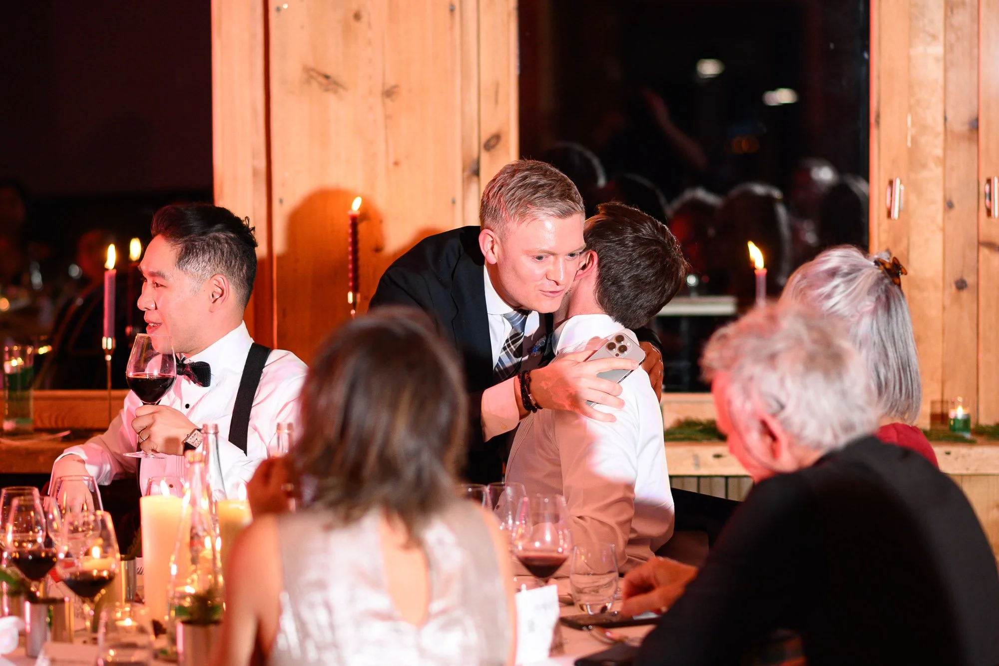 Des personnes assises à une table lors d'une célébration ou d'un dîner, avec un homme en costume qui embrasse ou parle à un autre jeune homme, dans une ambiance chaleureuse avec bougies et décorations en bois.
