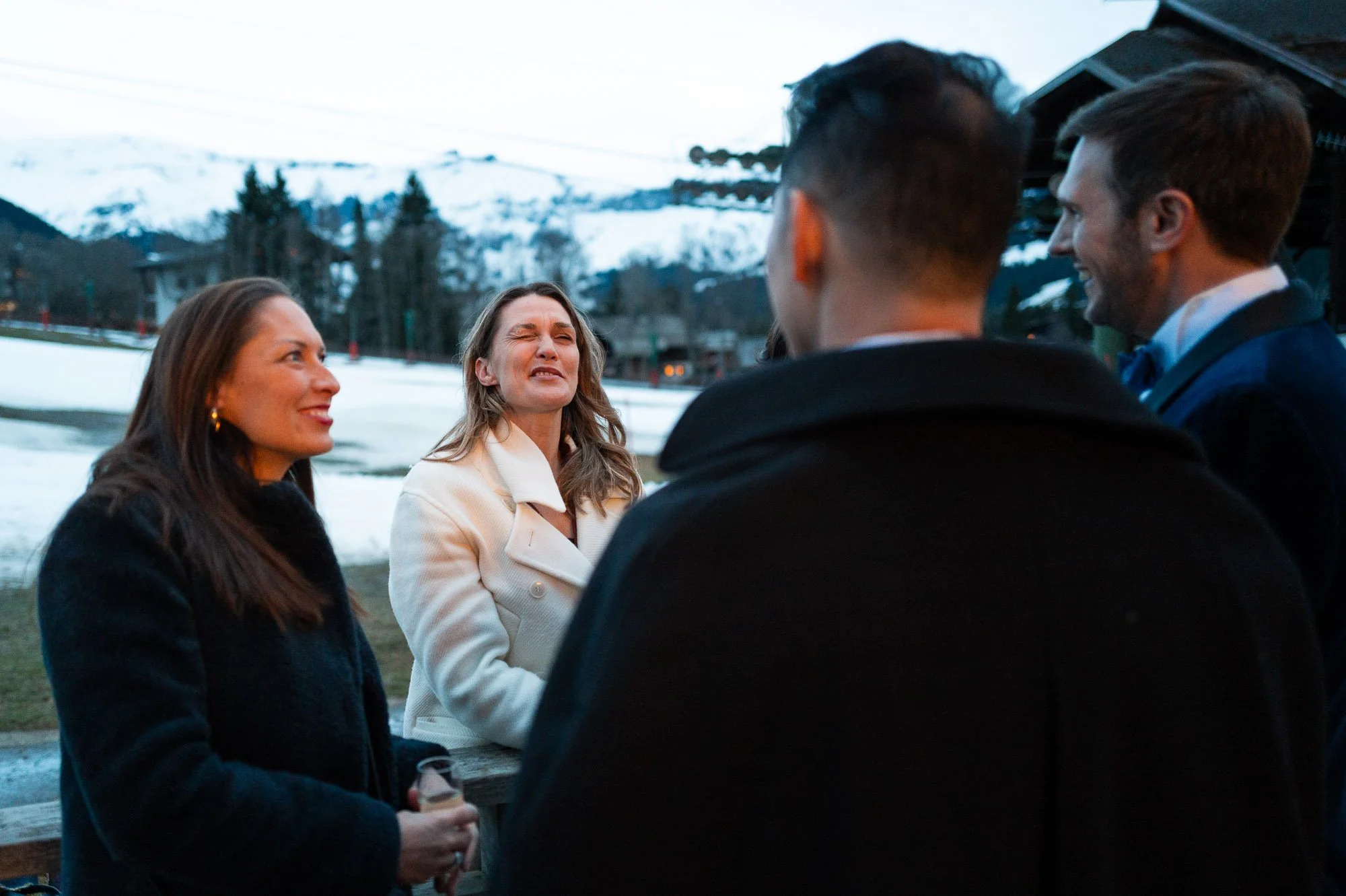 Groupe de quatre personnes discutant en extérieur dans un cadre hivernal avec neige et montagnes en arrière-plan, deux femmes et deux hommes. La femme à gauche tient un verre de boisson.