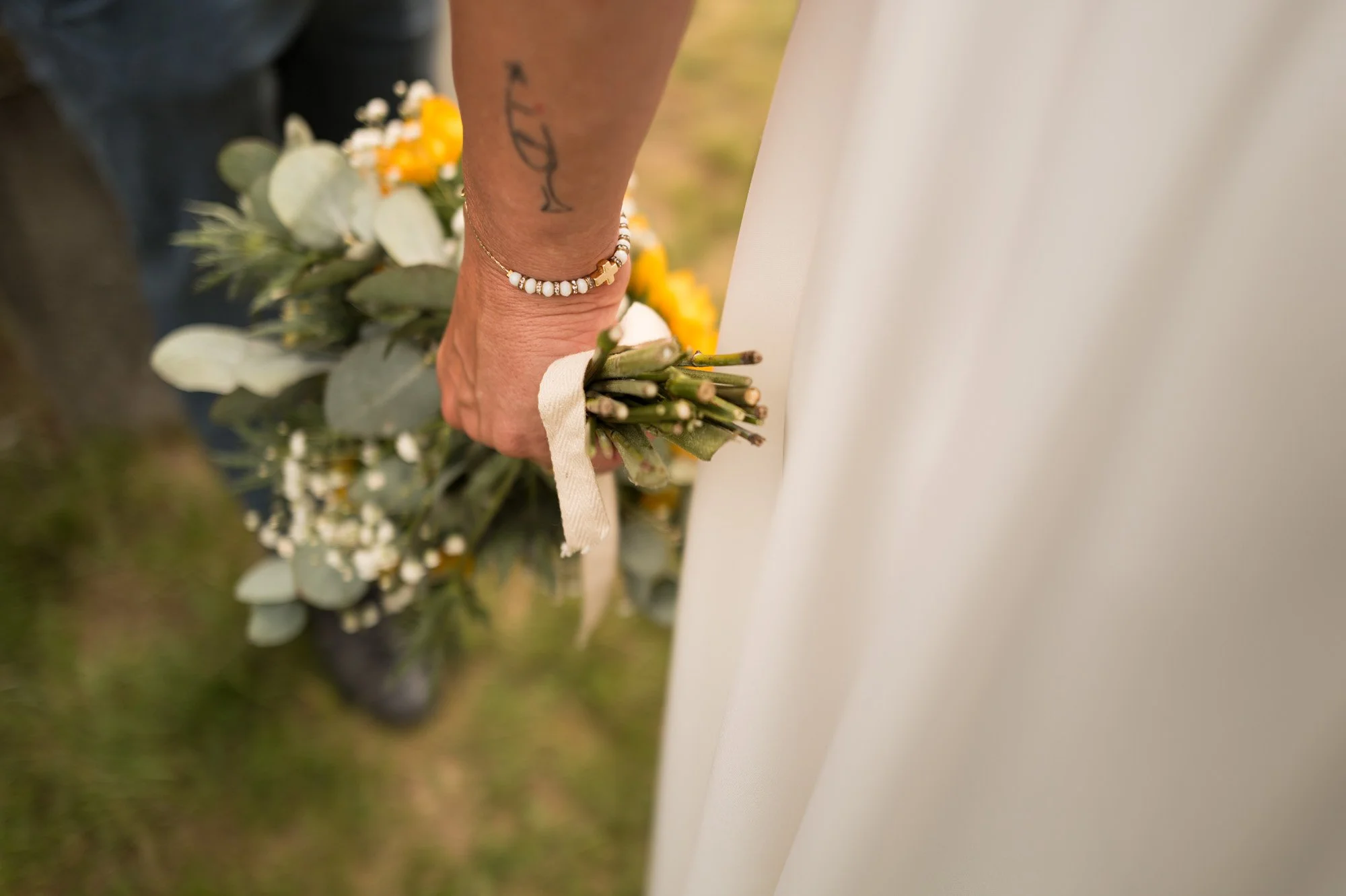 Une personne tient un bouquet de fleurs près d'une robe de mariage, portant un bracelet en perles et un tatouage sur le bras.