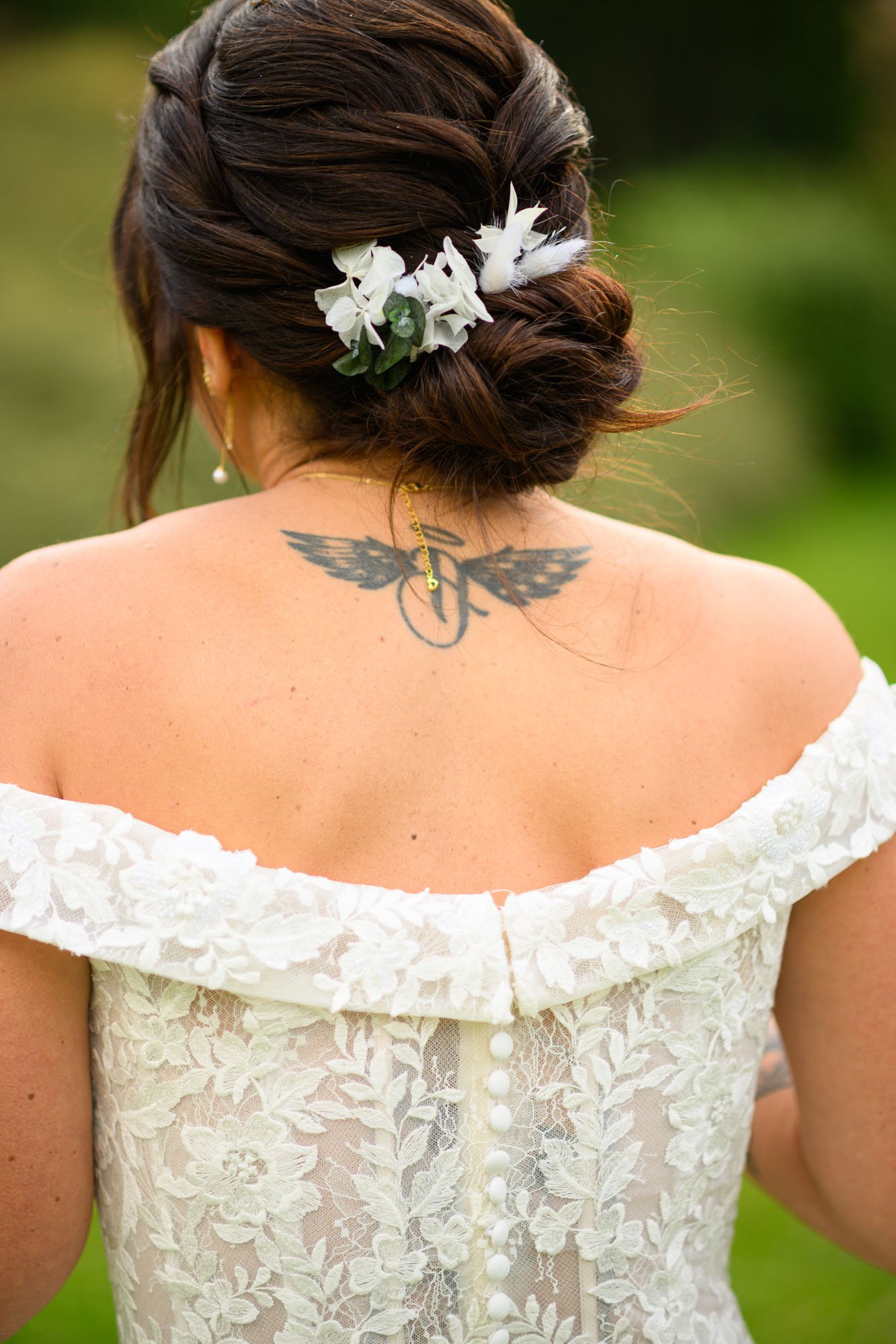 Femme vue de dos avec une coiffure brune tressée, portant des boucles d'oreilles avec perles, une robe blanche en dentelle à épaules dénudées, et un tatouage d'ailes avec un cercle sur le haut du dos.
