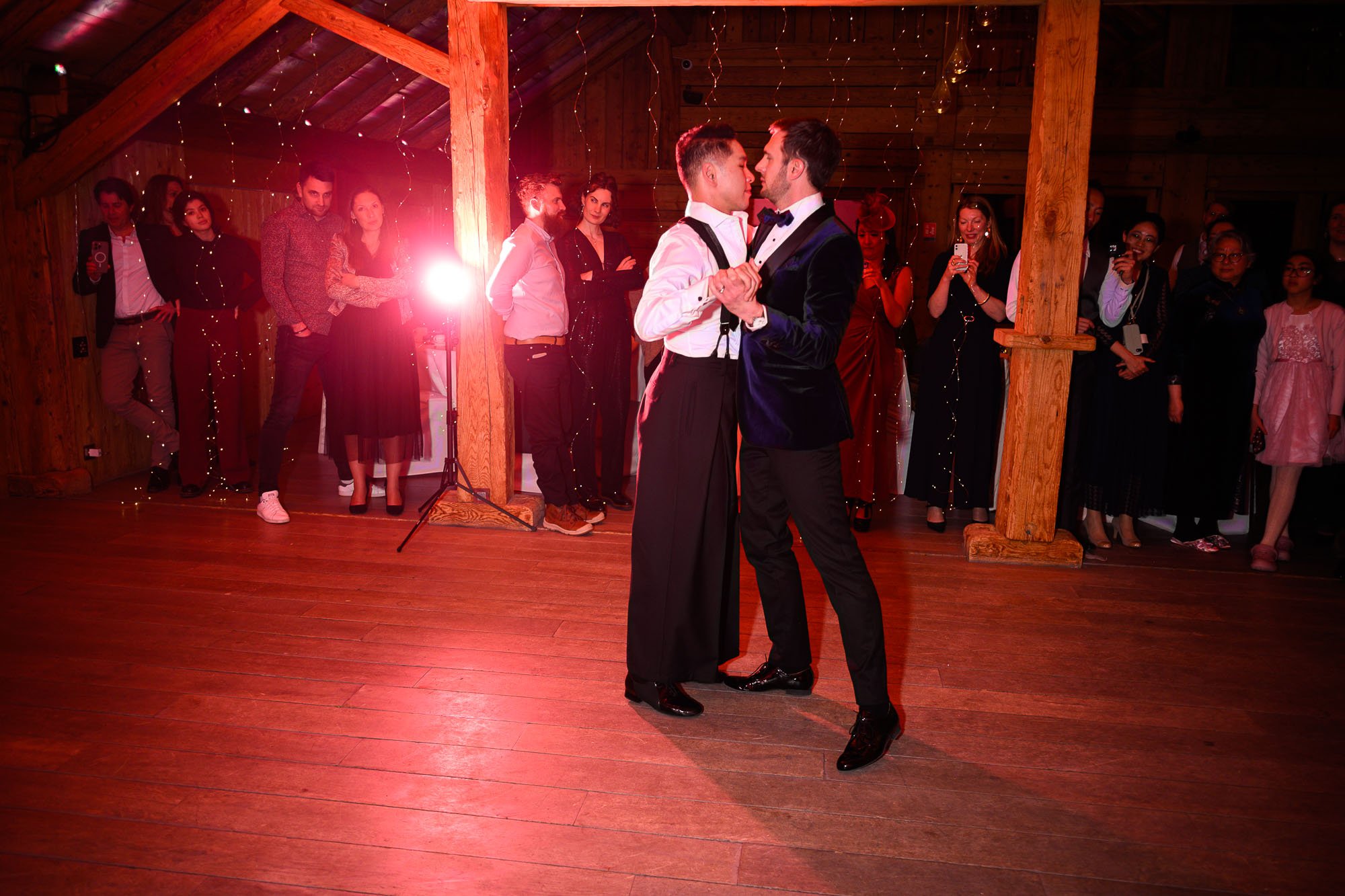 Deux hommes dansent ensemble, l'un en costume noir avec une chemise blanche et l'autre en queues de pie, lors d'une cérémonie ou d'une fête, entourés de spectateurs dans un décor en bois avec des lumières.