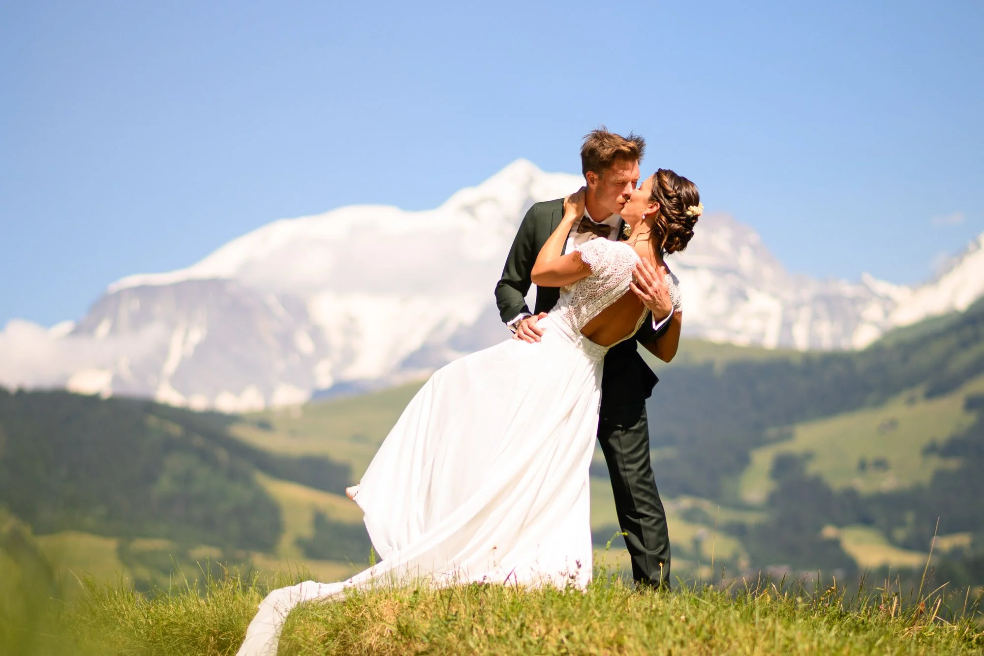 Un couple de mariés en robe blanche et costume noir s'embrassant dans un champ avec des montagnes enneigées en arrière-plan.