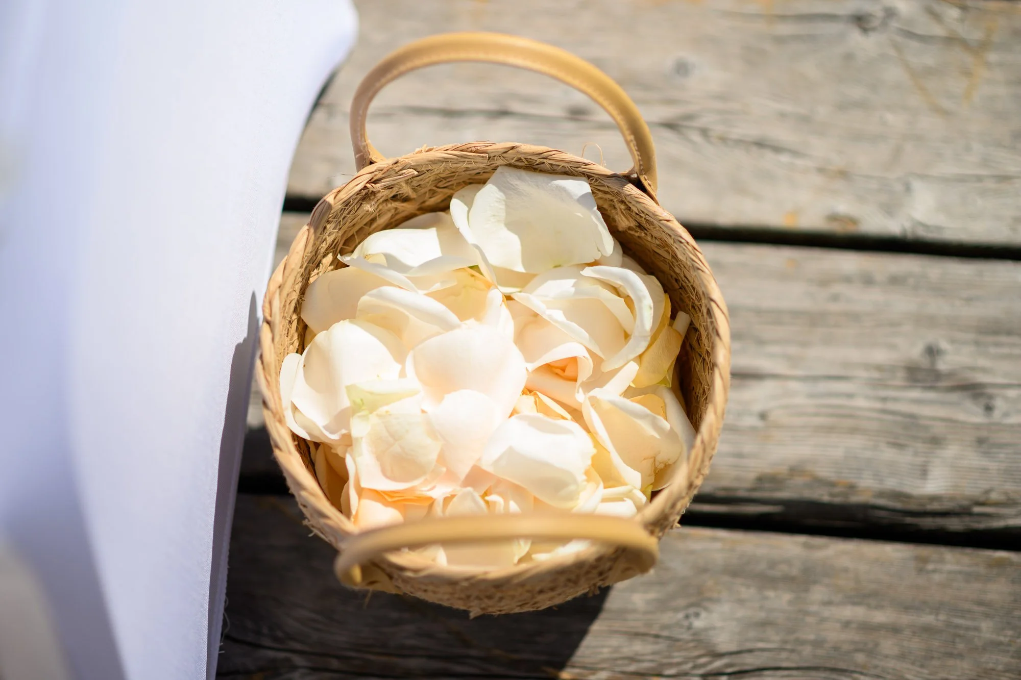 Un panier en osier rempli de pétales de roses blanches et crème, posé sur un pont en bois, avec une surface blanche à côté.