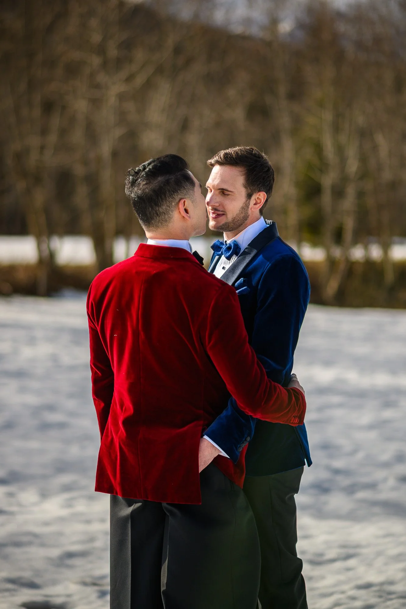 Deux hommes en costumes élégants s'embrassent dehors, avec un fond d'arbres et de neige.