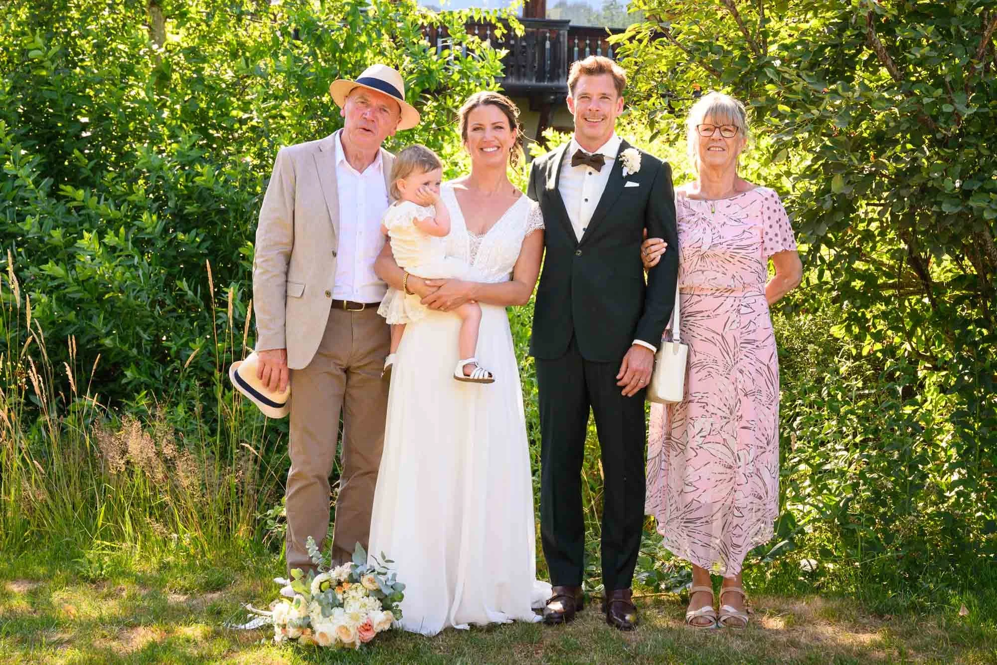 Famille réunie lors d'un mariage en plein air, avec un couple de jeunes mariés au centre, entourés de membres de leur famille, sous un ciel ensoleillé et un arbre en arrière-plan.