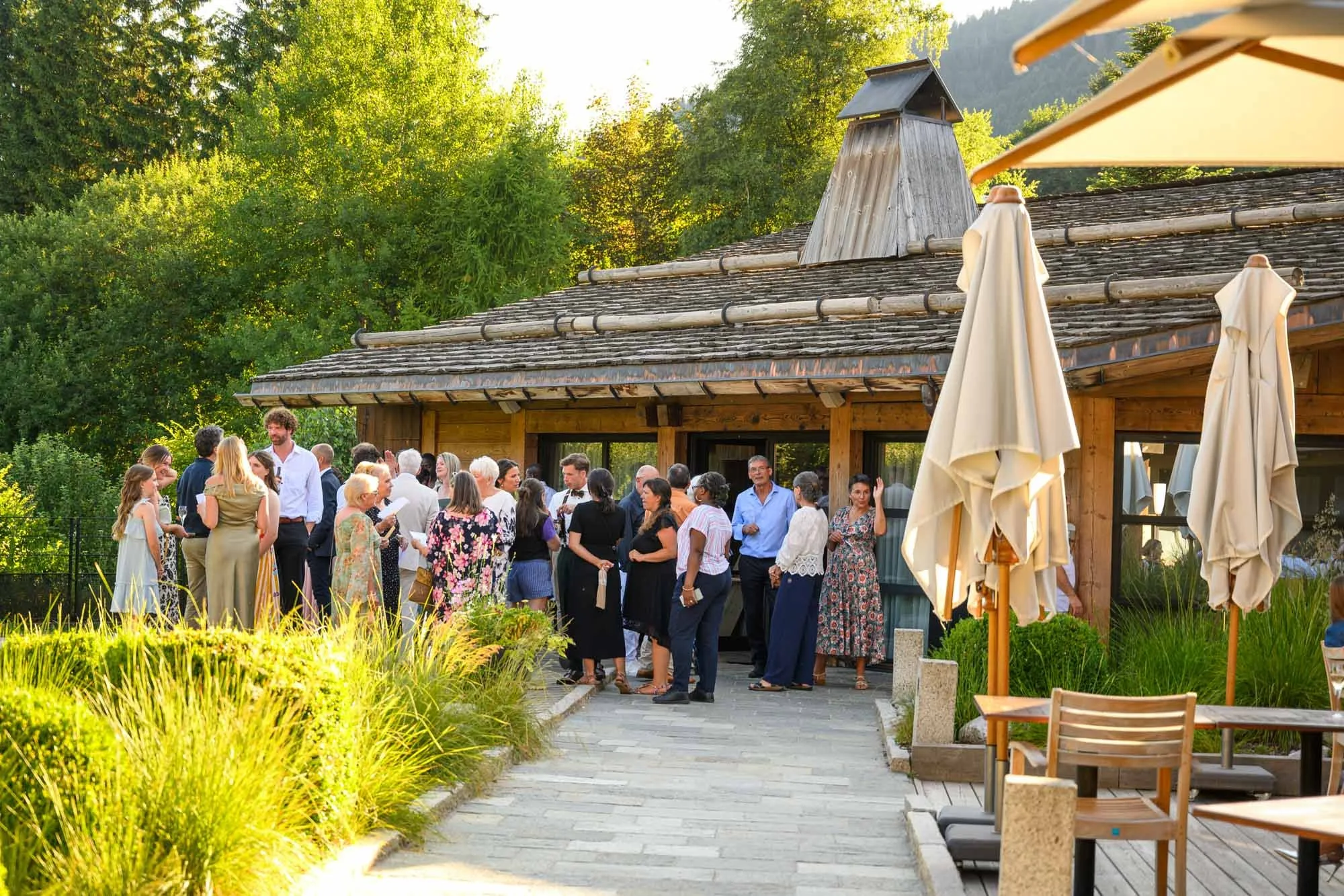 Groupe de personnes réunies à l'extérieur devant un bâtiment en bois lors d'une fête ou d'un événement social, avec des parasols fermés, dans un environnement verdoyant et ensoleillé.