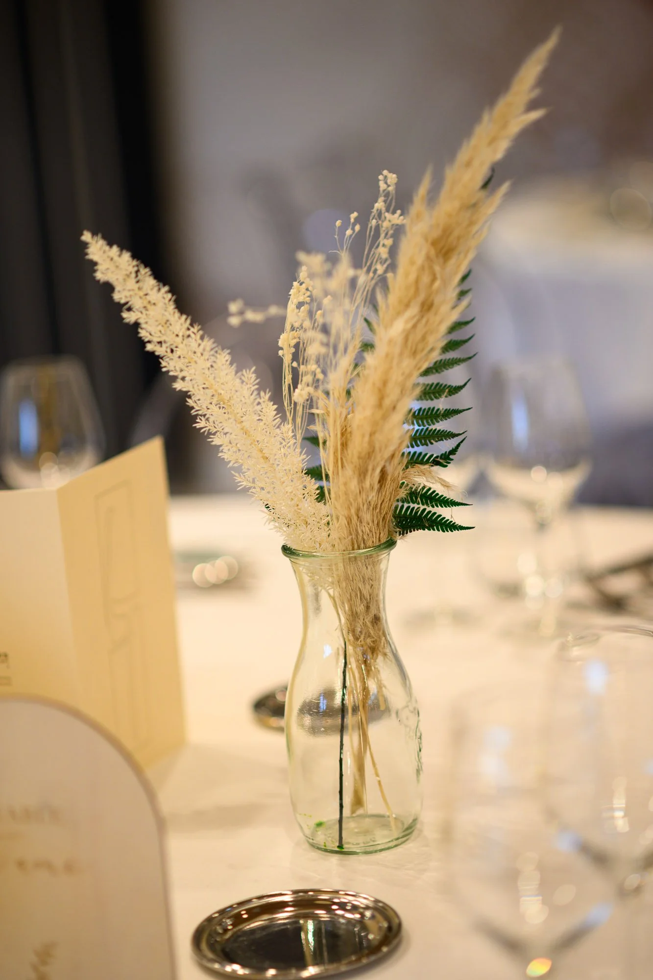 Vase en verre avec des fleurs séchées beige et des feuilles vertes, posé sur une table pour un événement.