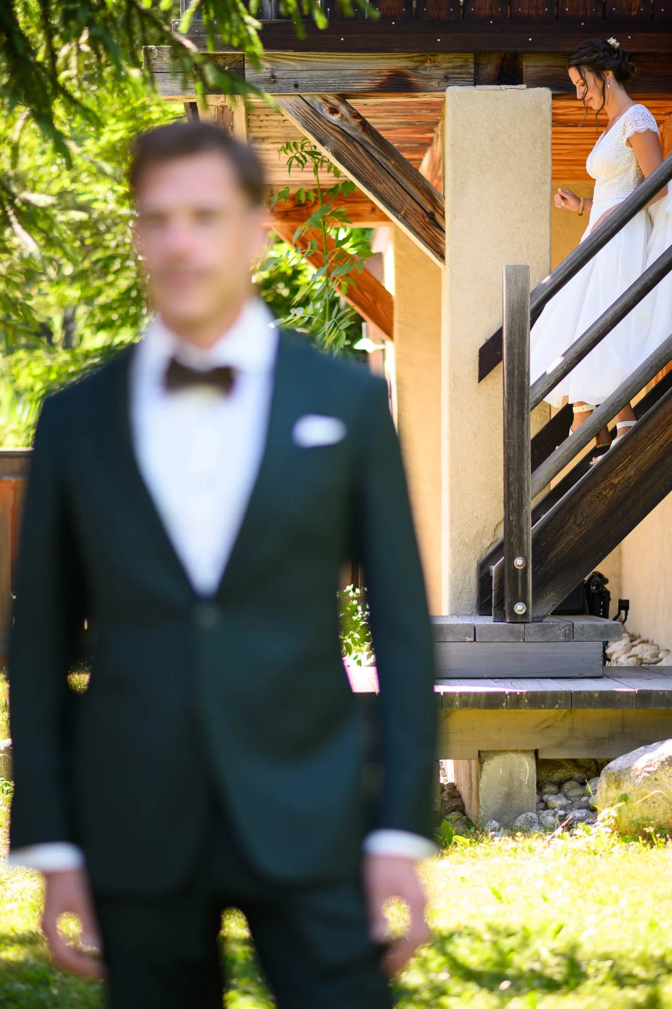 Une femme en robe blanche descend un escalier en bois, à l'extérieur, pendant qu'un homme en costume sombre, flou dans le premier plan, attend ou la regarde.