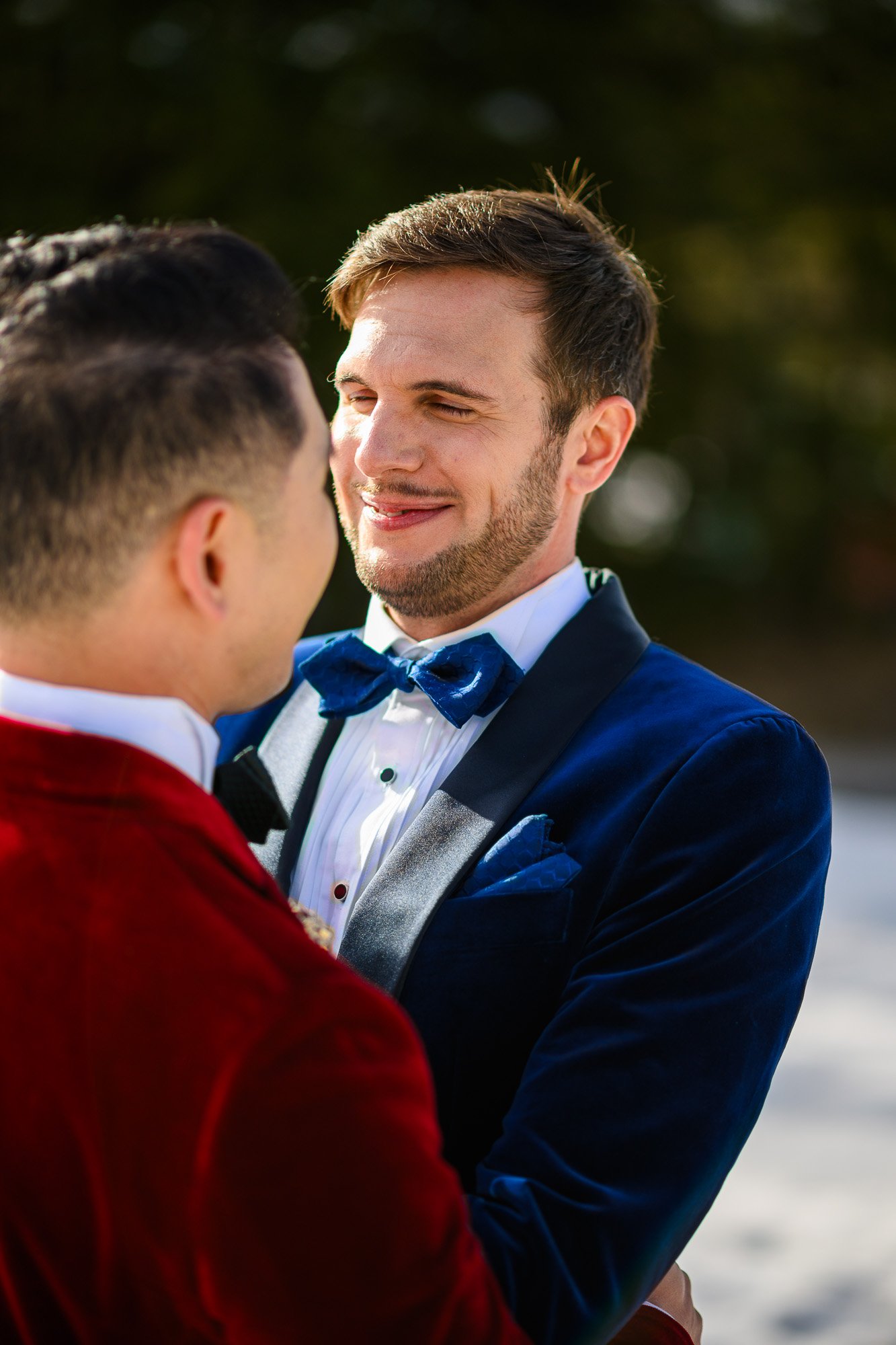Deux hommes en costume habillé, l'un avec un nœud papillon bleu, s'enlacent et se regardent tendrement, lors d'une cérémonie ou événement en plein air.