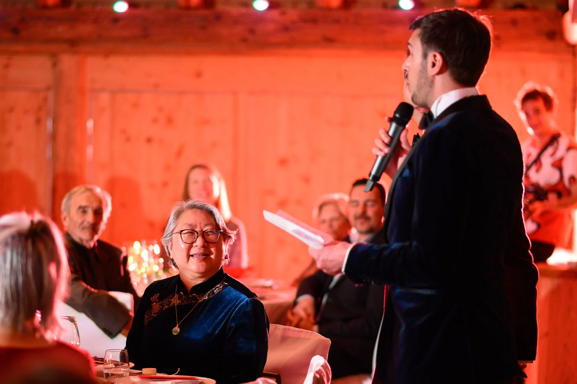 Un homme en tuxedo donne un discours avec un microphone lors d'une réception ou d'un dîner formel, à destination d'un groupe de personnes assises à une table, dans une salle en bois chaleureuse.
