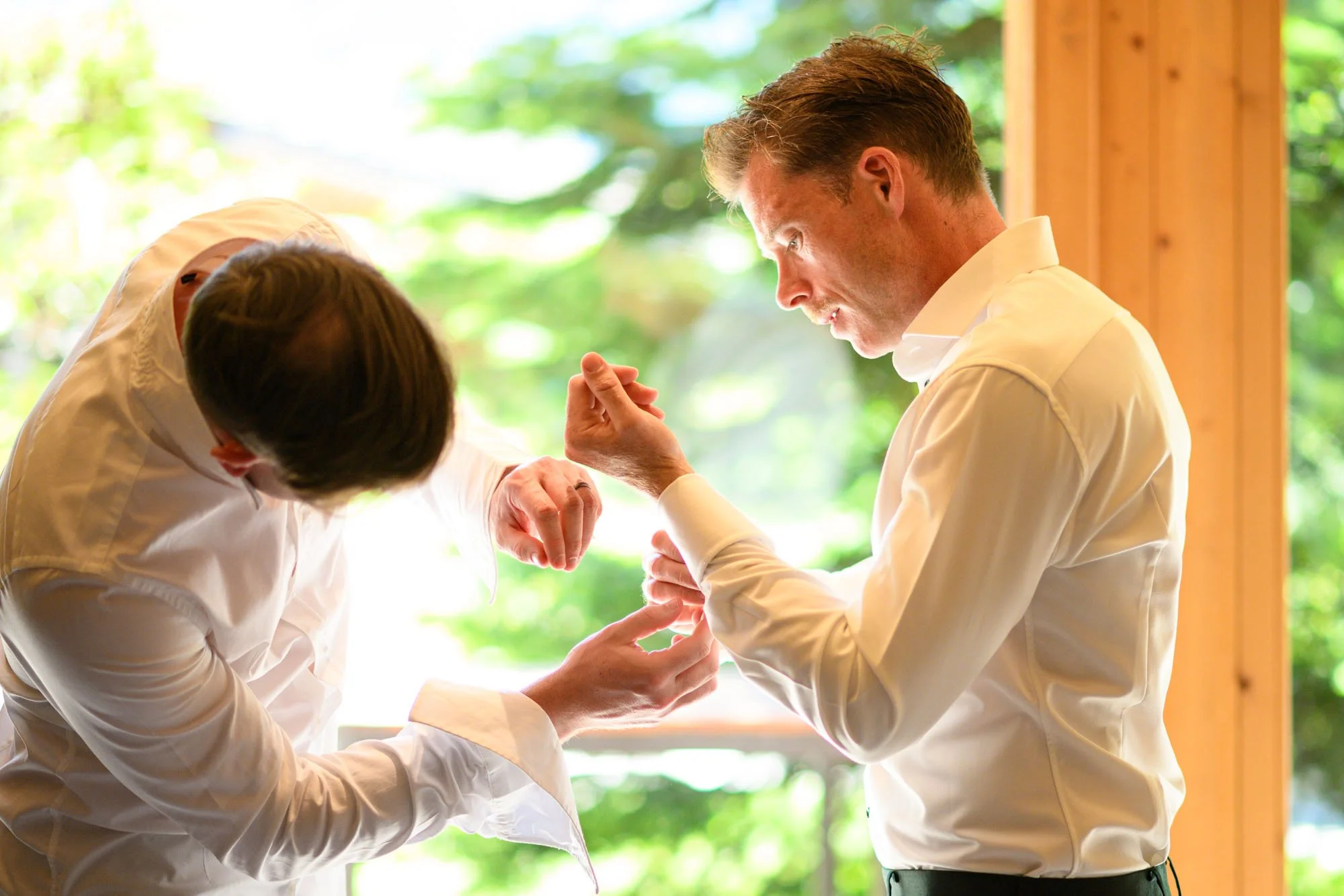 Deux hommes habillés en chemises blanches, en train de s'ajuster les boutons de manchette dans une pièce avec beaucoup de lumière naturelle et un décor en bois, avec un fond de verdure floue visible par la fenêtre.