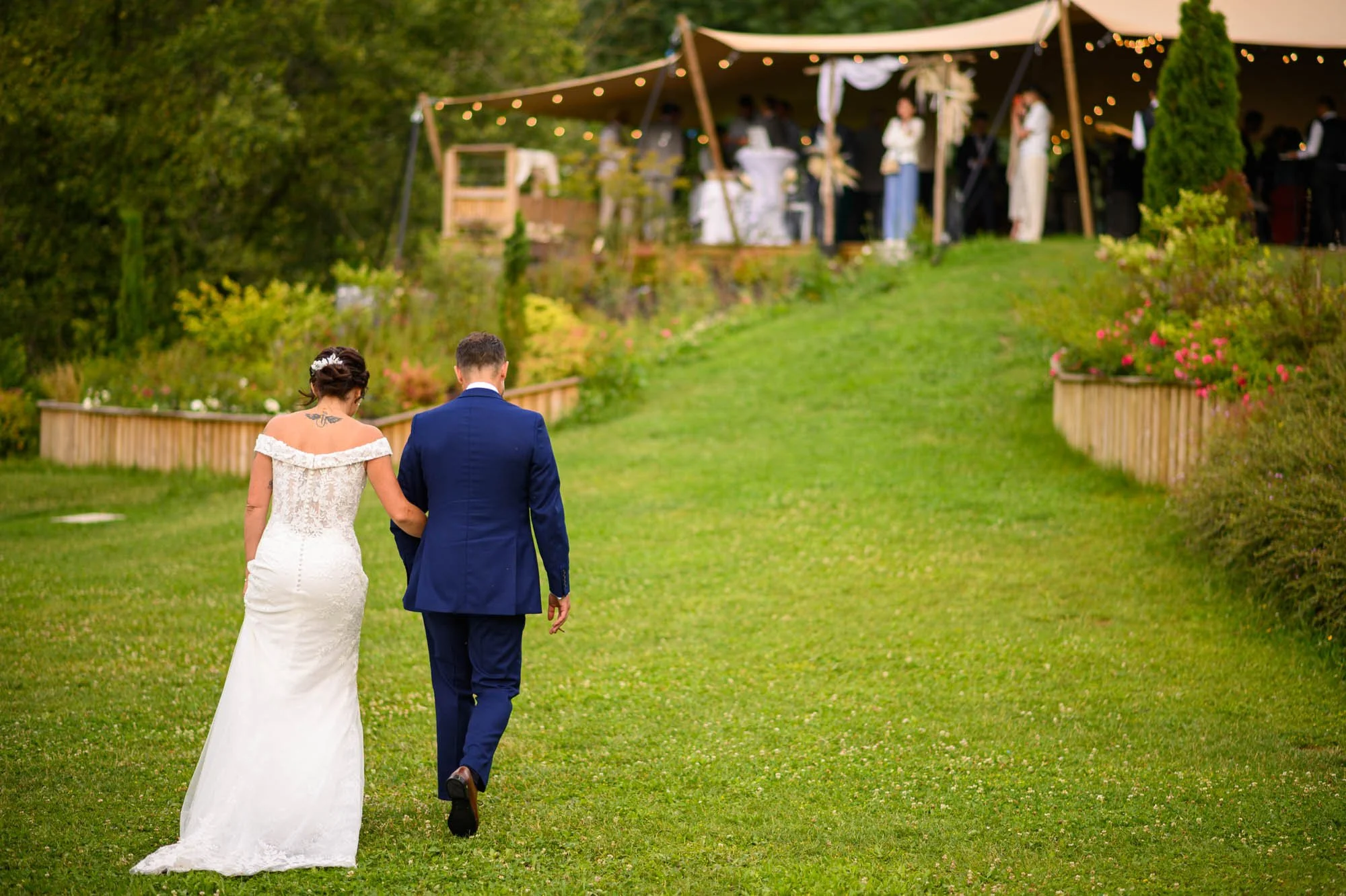 Un couple marche dans un jardin lors d'un mariage en extérieur, avec une tente décorée de guirlandes lumineuses en arrière-plan.