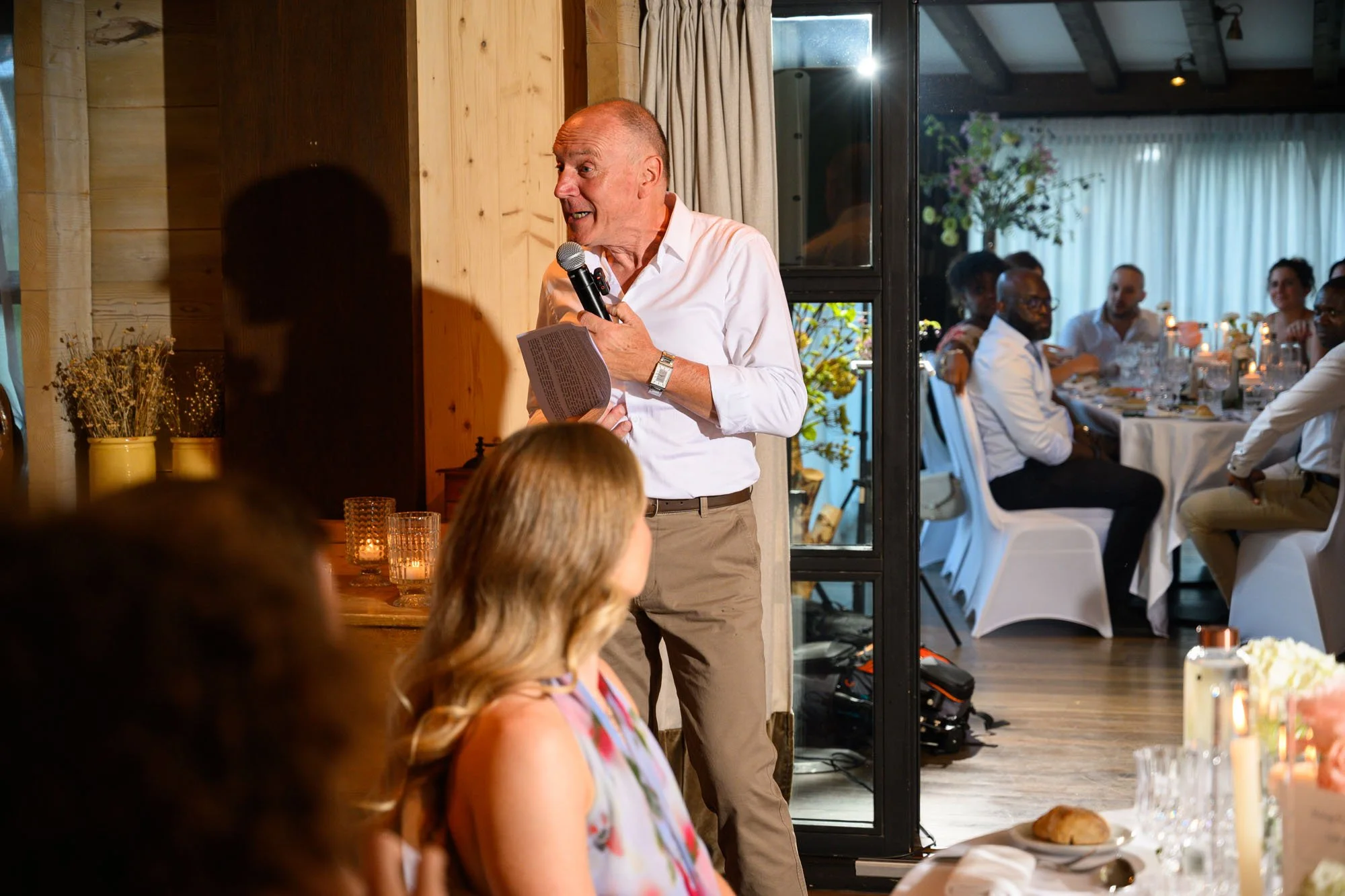 Un homme âgé en chemise blanche parlant avec un micro lors d'un événement avec un groupe de personnes assises à une table dans une salle de réception