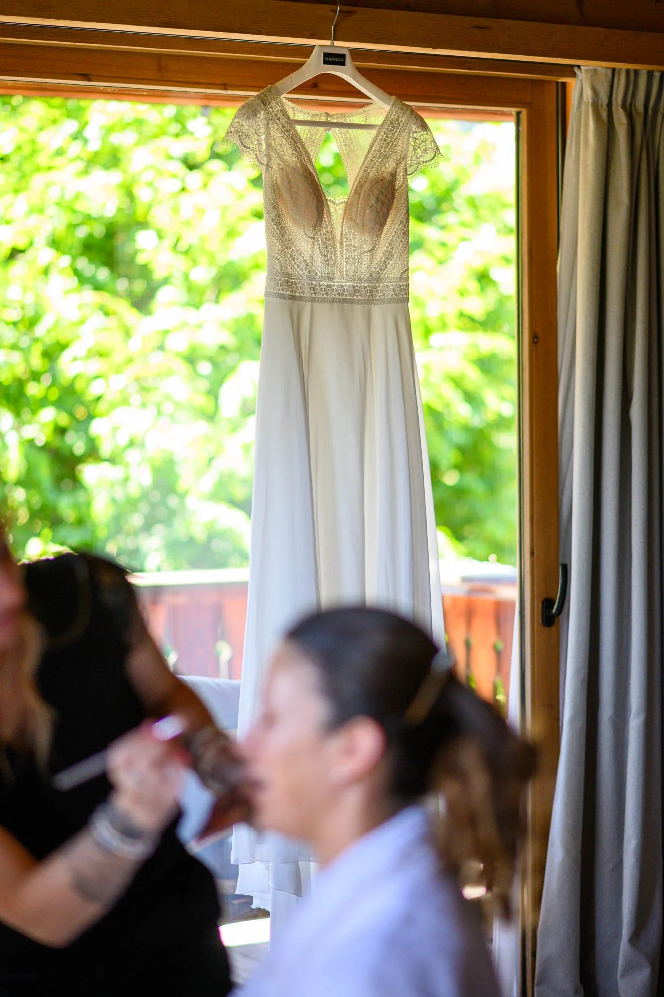 Une robe de mariage blanche suspendue à un cintre en bois dans une pièce avec une fenêtre sur un fond lumineux de feuillage vert.