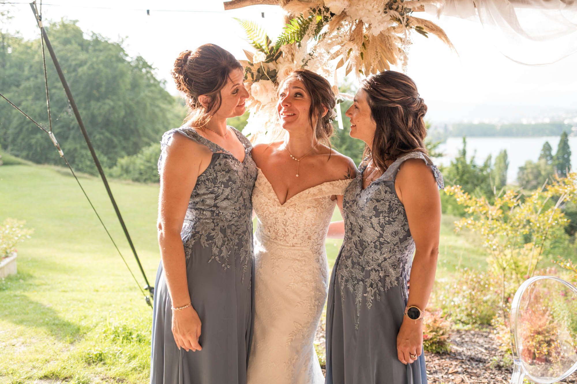 Trois femmes habillées pour un mariage, dont une en robe de mariée blanche et deux en robes gris avec des détails floraux, se regardant et souriant sous un décor extérieur avec des fleurs et la nature en arrière-plan.