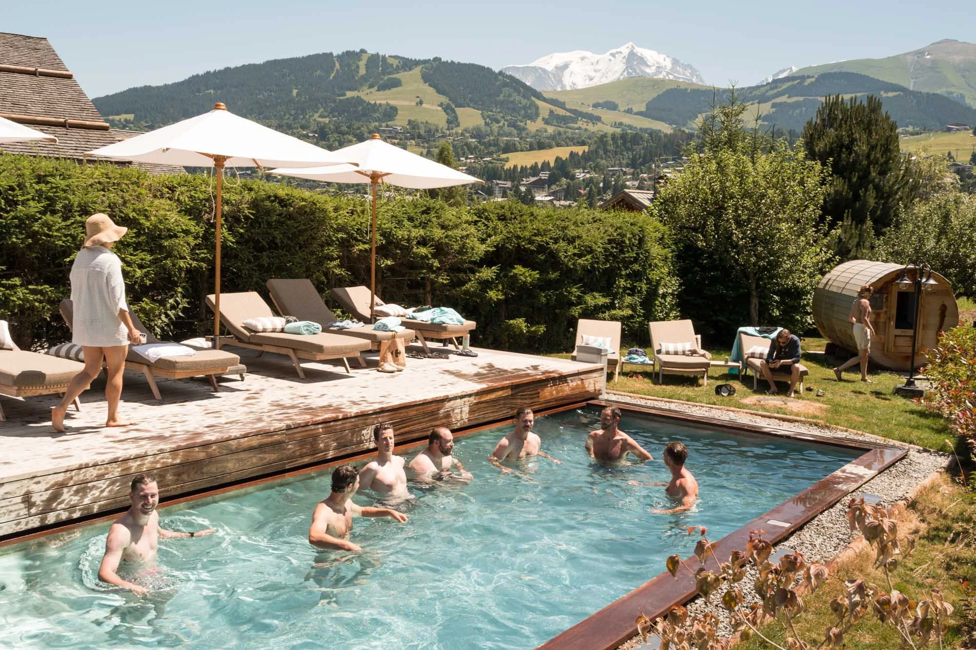 Groupe de personnes profitant d'une baignade dans une piscine extérieure entourée de chaises longues, parasols et un sauna en bois dans un paysage montagnard ensoleillé.