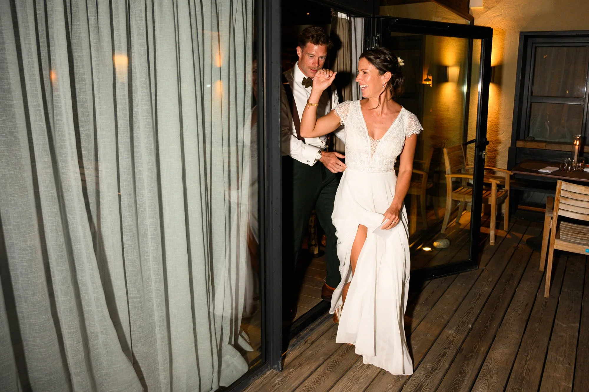 Un couple marié sortant d'une pièce lors d'un mariage, la mariée en robe blanche avec une fente, l'homme en costume tuxedo, ils sourient et semblent joyeux.