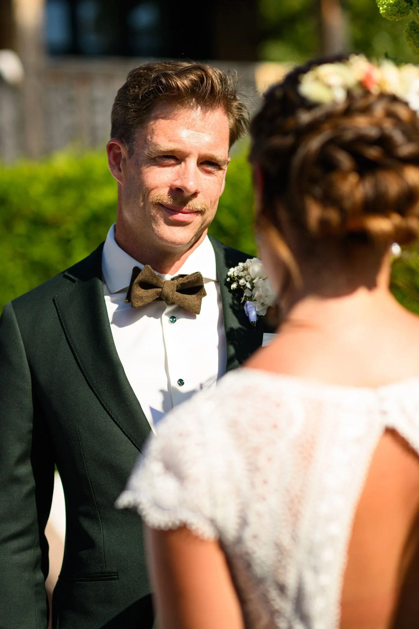 Un homme en costume de mariage noir avec un nœud papillon marron sourit, regardant une femme dont le dos est visible, lors d'une cérémonie en plein air, avec un fond vert et ensoleillé.