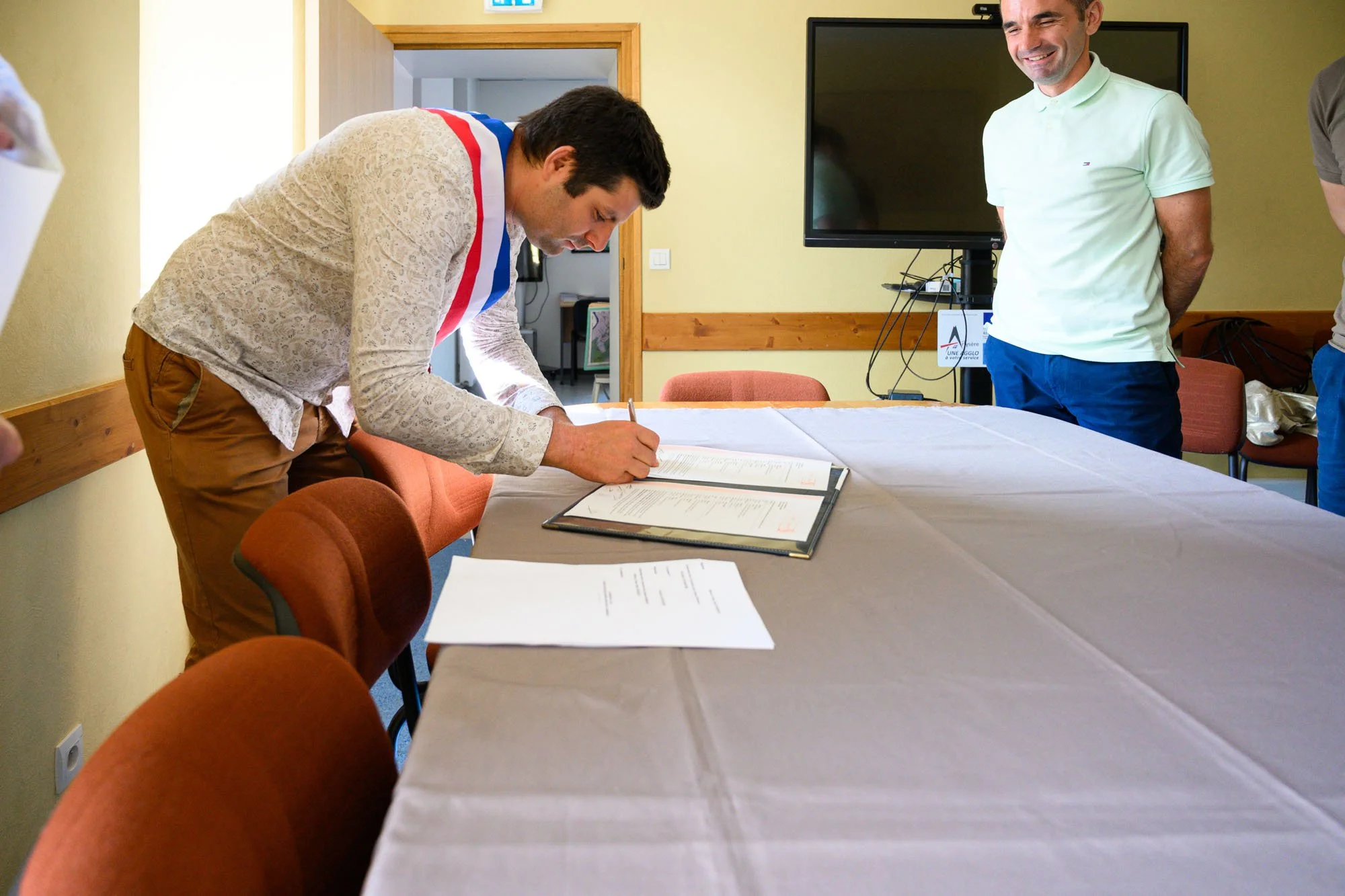 Un homme appose sa signature sur un document officiel sur une table, d'autres personnes sont présentes dans la pièce, un écran télé est visible à l'arrière.