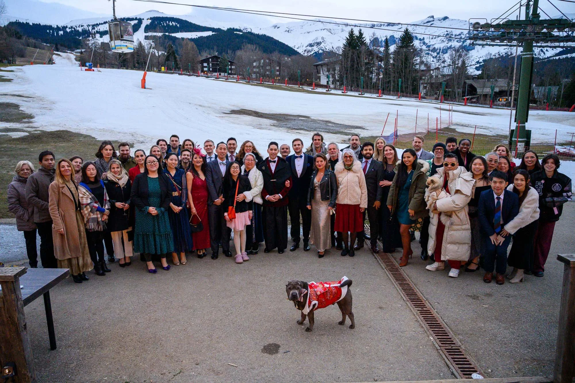 Groupe de personnes rassemblées devant une station de ski avec montagnes enneigées en arrière-plan. Un chien déguisé en Père Noël est en premier plan.