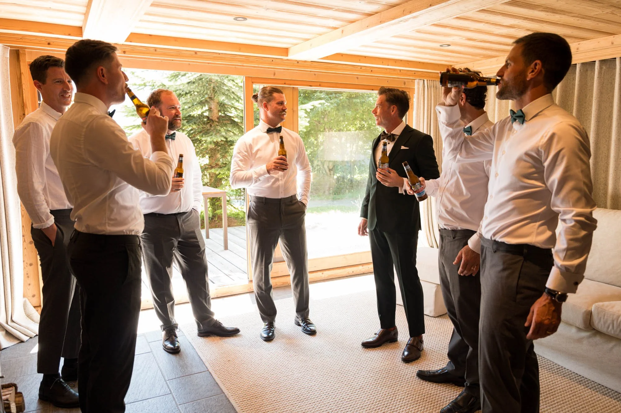 Groupe d'hommes en costume avec des chemises blanches et des nœuds papillons, buvant de la bière dans une pièce lumineuse avec grandes fenêtres donnant sur un jardin.
