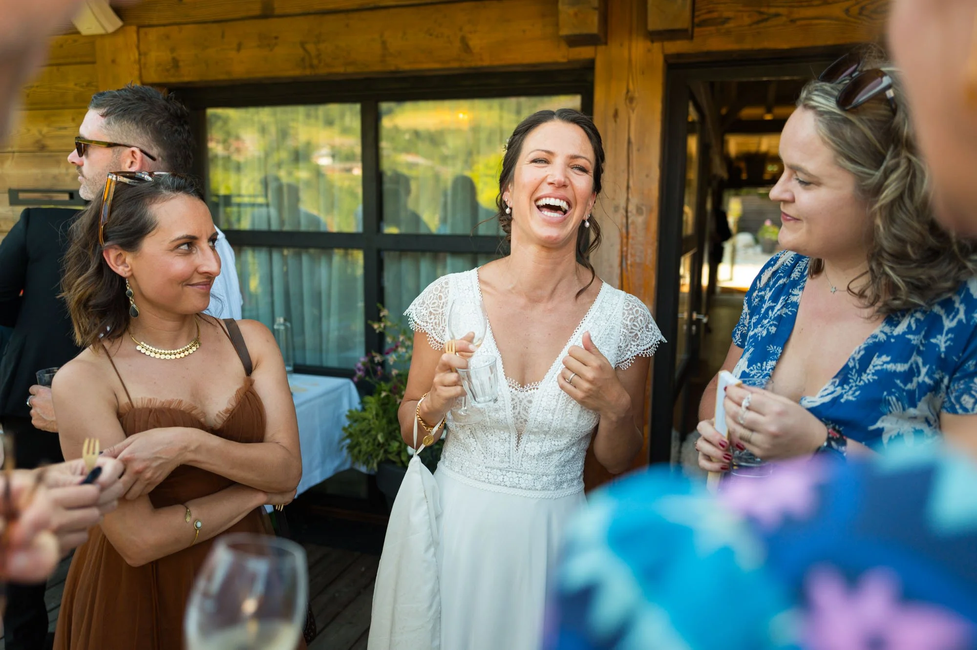 Une femme souriante en robe blanche parle à un groupe de personnes lors d'une fête en intérieur, avec un décor en bois.