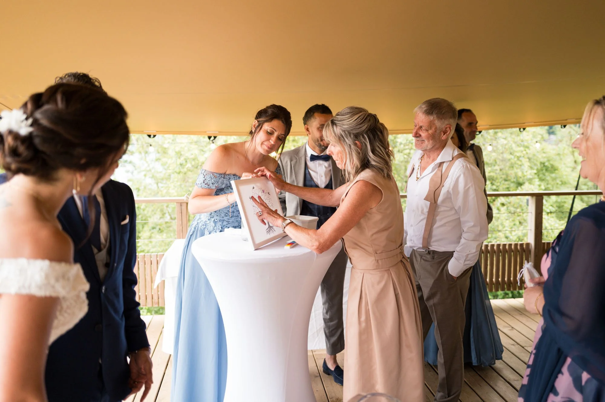 Groupe de personnes lors d'un mariage, se réunissant autour d'une table, échangeant des cadeaux ou des souvenirs, avec un décor extérieur lumineux et verdoyant.