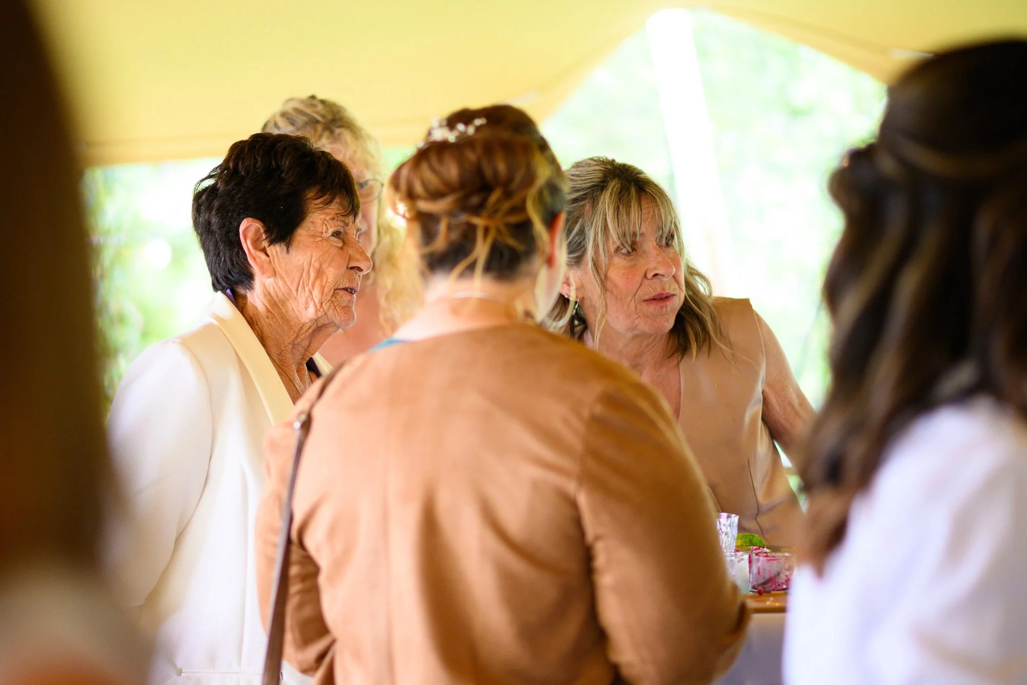 Un groupe de femmes âgées discutant ensemble lors d'un événement en intérieur, sous une tente, avec une lumière naturelle en arrière-plan.