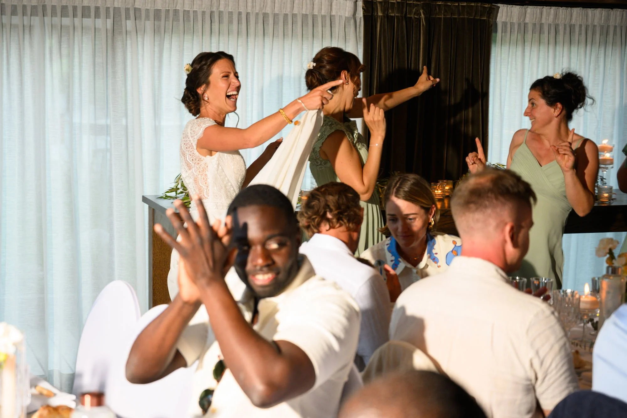 Groupe de personnes à une fête, certaines dansant et riant, devant une fenêtre avec rideaux blancs, ambiance festive avec bougies.