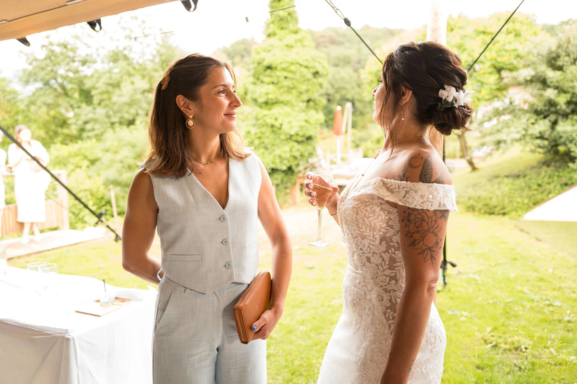 Deux femmes en robe de mariage bavardent lors d'une réception en plein air, avec un décor verdoyant en arrière-plan.