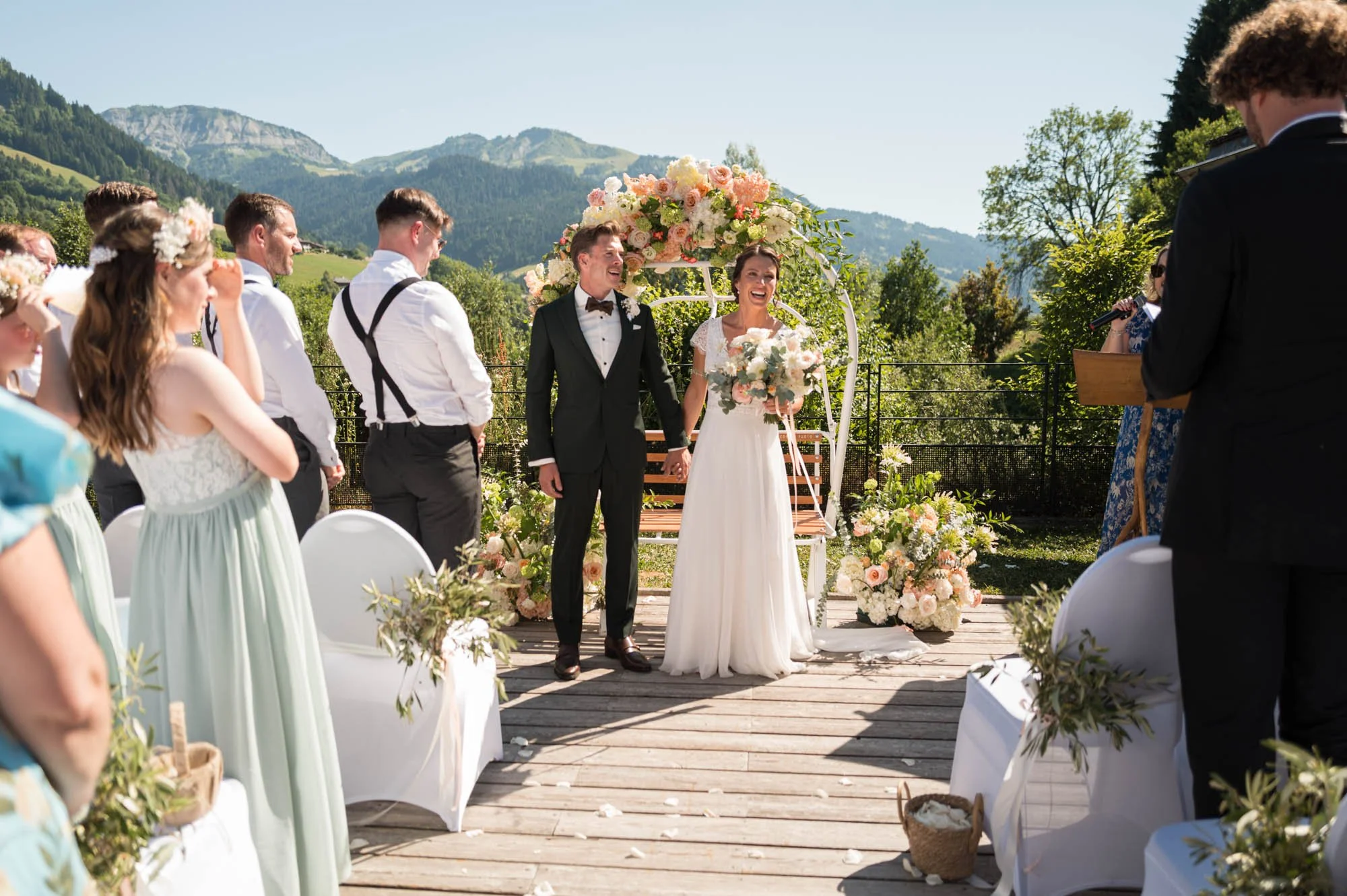 Un couple marié posant pour la photo lors de leur mariage en plein air, entouré d'amis et famille dans un paysage montagneux.