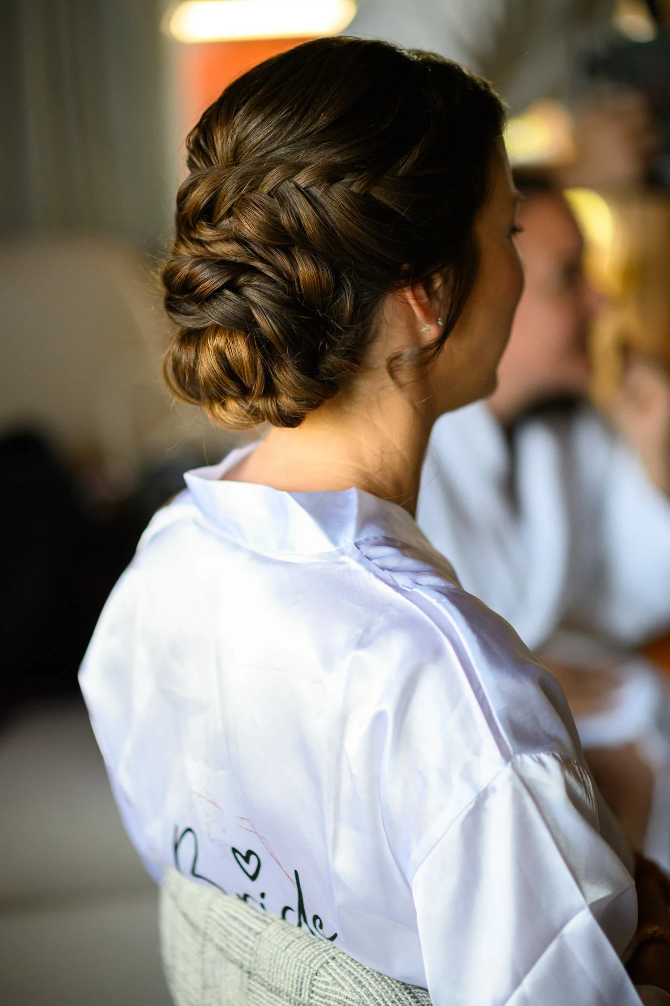 Une femme avec une coiffure en chignon tressé, portant une blouse blanche, assise de profil, en intérieur.