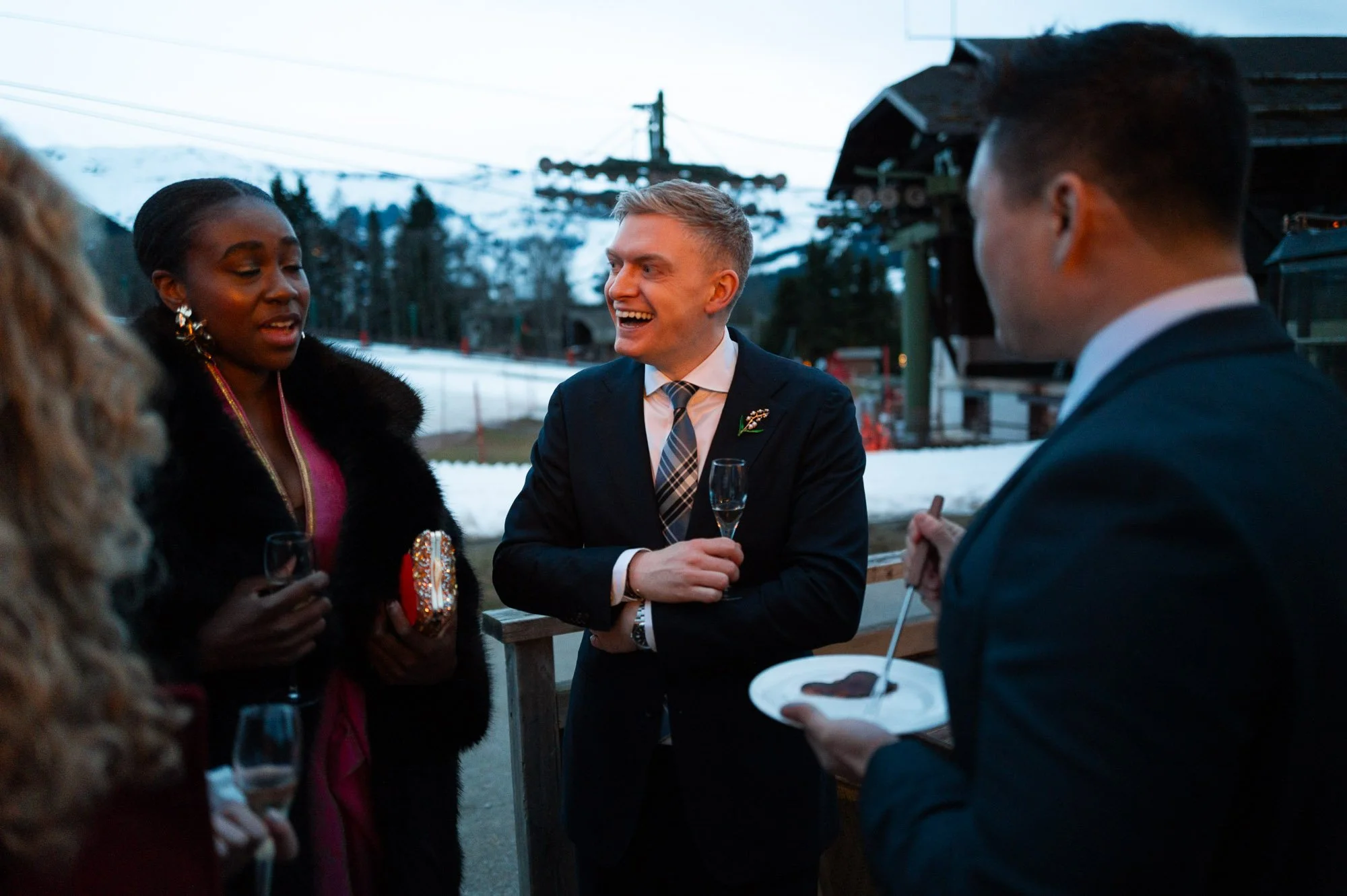Groupe de quatre personnes en costume lors d'une fête en extérieur dans un environnement enneigé, avec montagnes en arrière-plan, discutant et riant.