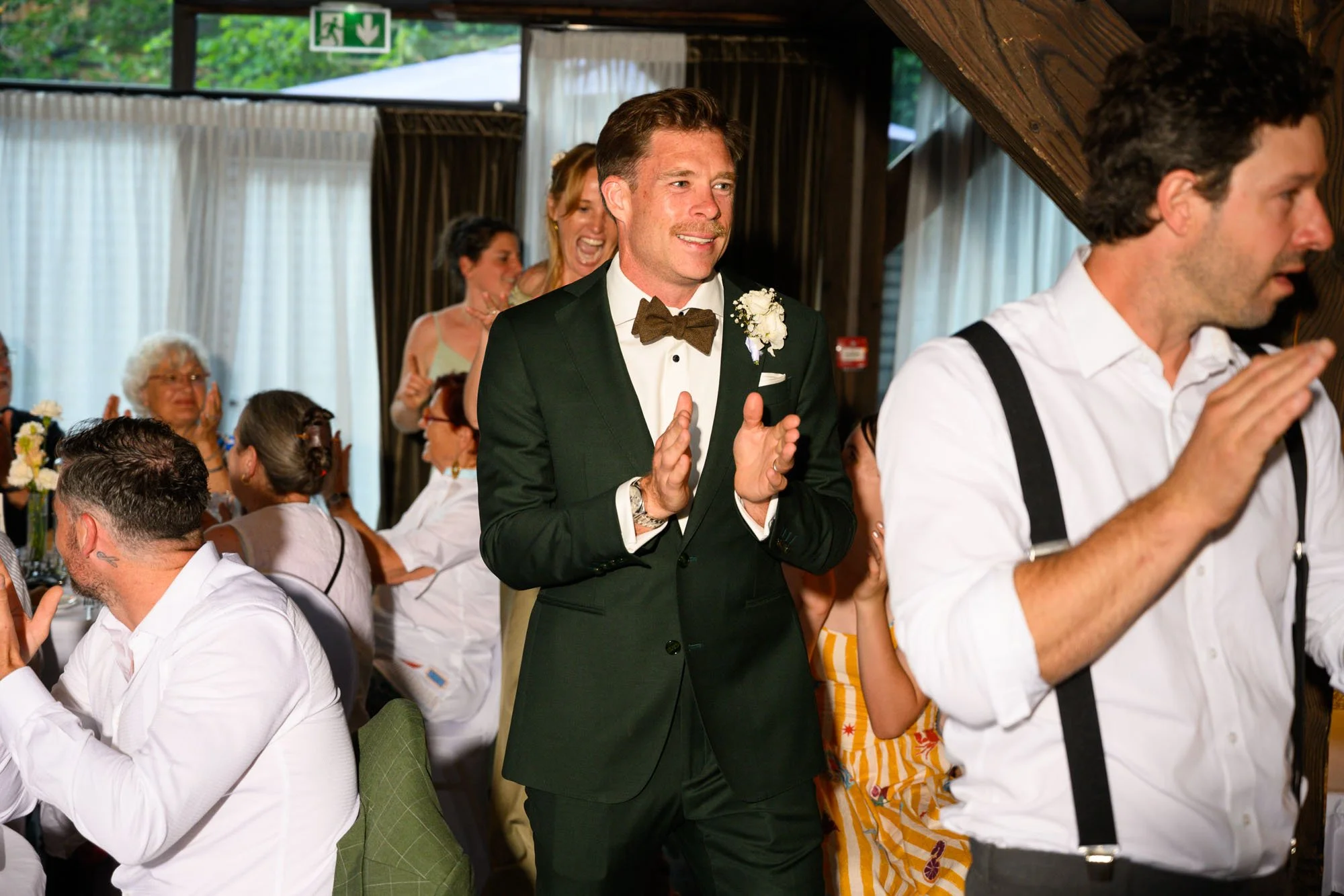 Un groupe de personnes à une célébration, probablement un mariage, dans une salle décorée, avec un homme en costume vert et nœud papillon au centre qui applaudit, entouré de personnes qui applaudent et sourient.