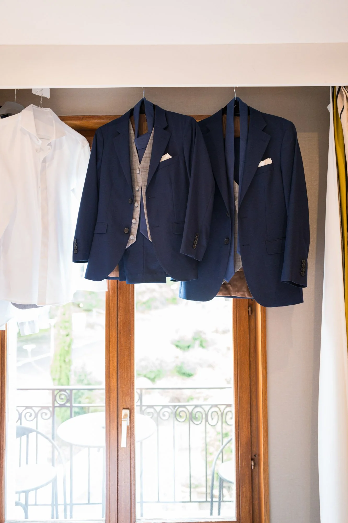 Costumes de mariage à l'intérieur, comprenant deux costumes bleus avec doublure grise, suspendus devant une fenêtre en bois avec vue sur un balcon en fer forgé.